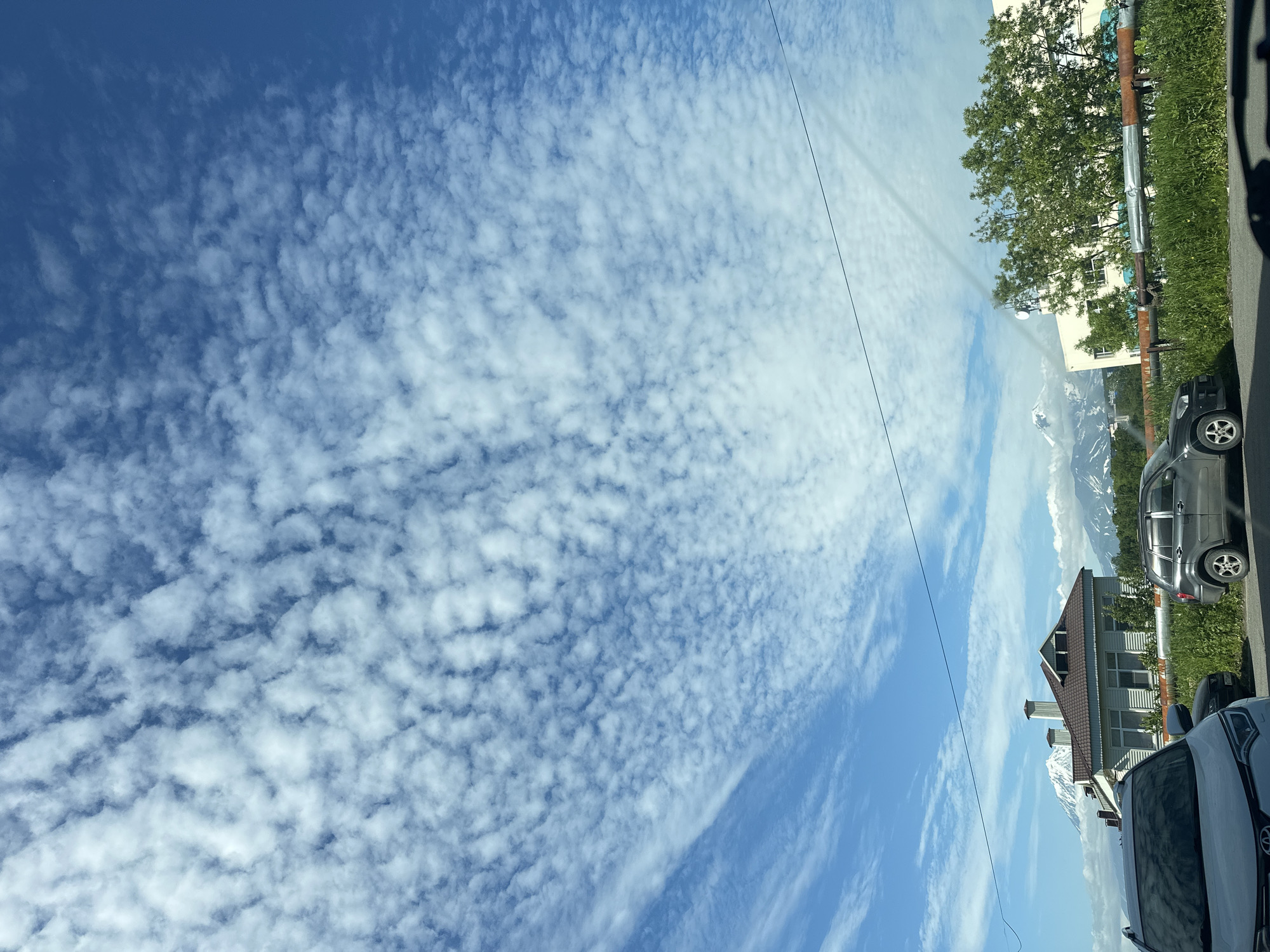 The image captures a cloudy sky over Petropavlovsk-Kamchatsky, Russia. In the foreground, several cars are parked on a roadside, with a white SUV in the left and a darker SUV in the middle. Behind the cars, a house with a brown roof is visible, along with a building with a light-colored facade and green trees. In the distance, snow-capped mountains are seen, suggesting a mountainous landscape. A thin wire runs horizontally across the frame, dividing the sky and the landscape. The overall scene depicts a typical day in a city with a backdrop of natural mountainous scenery.