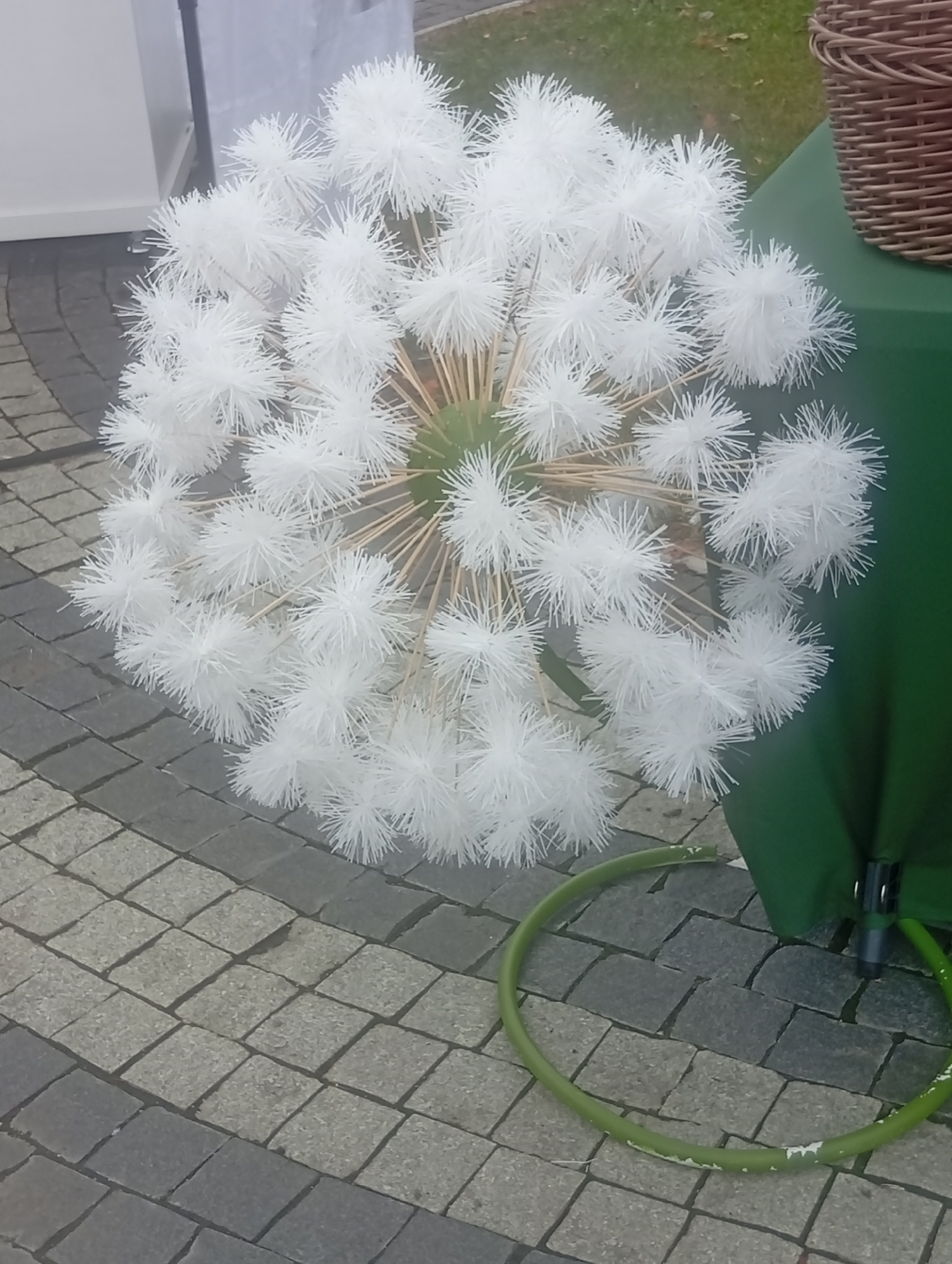 A large, artificial dandelion globe, constructed from white fluffy material attached to thin wooden sticks, is displayed outdoors on a grey and beige paved surface. The globe is positioned centrally in the frame, its fluffy seed-head components fanning outwards. Behind the dandelion, a dark brown woven basket is visible at the top right, resting on a dark green tablecloth that covers a table. A green hose lies in a circular shape on the pavement in the foreground. The background suggests an urban environment with a hint of grass and foliage. No people are present. The overall impression is of an outdoor decorative setup.