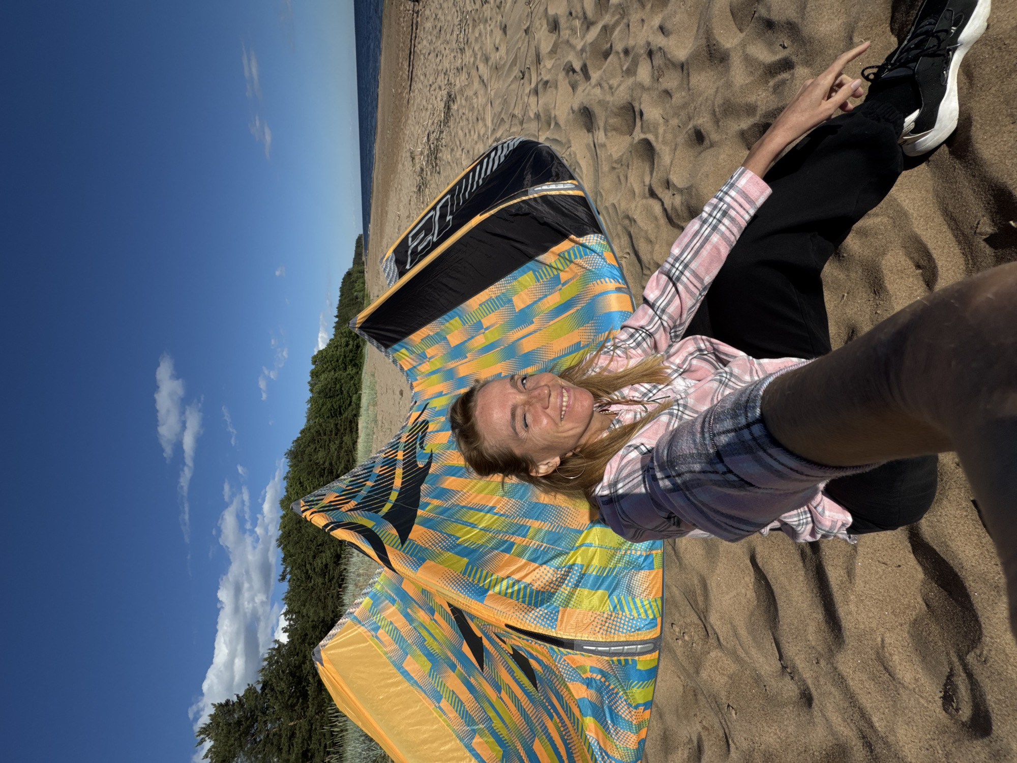 An adult female with blonde hair, wearing a pink and white plaid shirt and black pants, is sitting on a sandy beach with two colorful kites behind her. The kites are predominantly orange, blue, and yellow with black accents. She is looking at the camera and winking. The background shows a calm blue body of water, a sandy shore, and a dense green forest. The sky is clear with a few scattered white clouds. The location is identified as Zelenaya Roshcha, Russia.