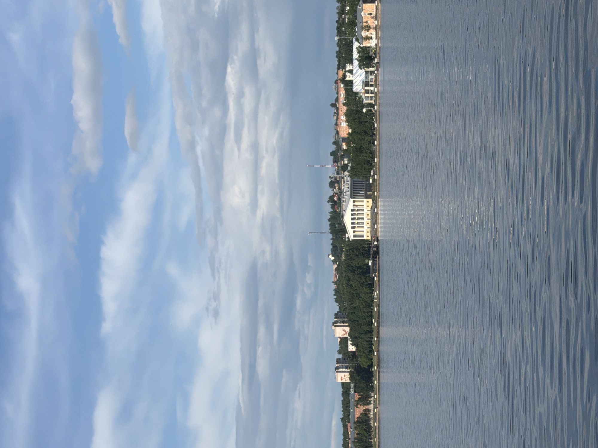 The image shows a view of the city of Votkinsk, Russia, from across the Votkinsk Reservoir. The horizon is oriented vertically with the sky and clouds on the left, the city center in the middle, and the reservoir on the right. Several buildings are visible in the city center, including taller apartment buildings and a few industrial structures with smokestacks. Trees line the shoreline. The water surface of the reservoir is slightly rippled. The sky is partly cloudy with streaks of white and light grey clouds.