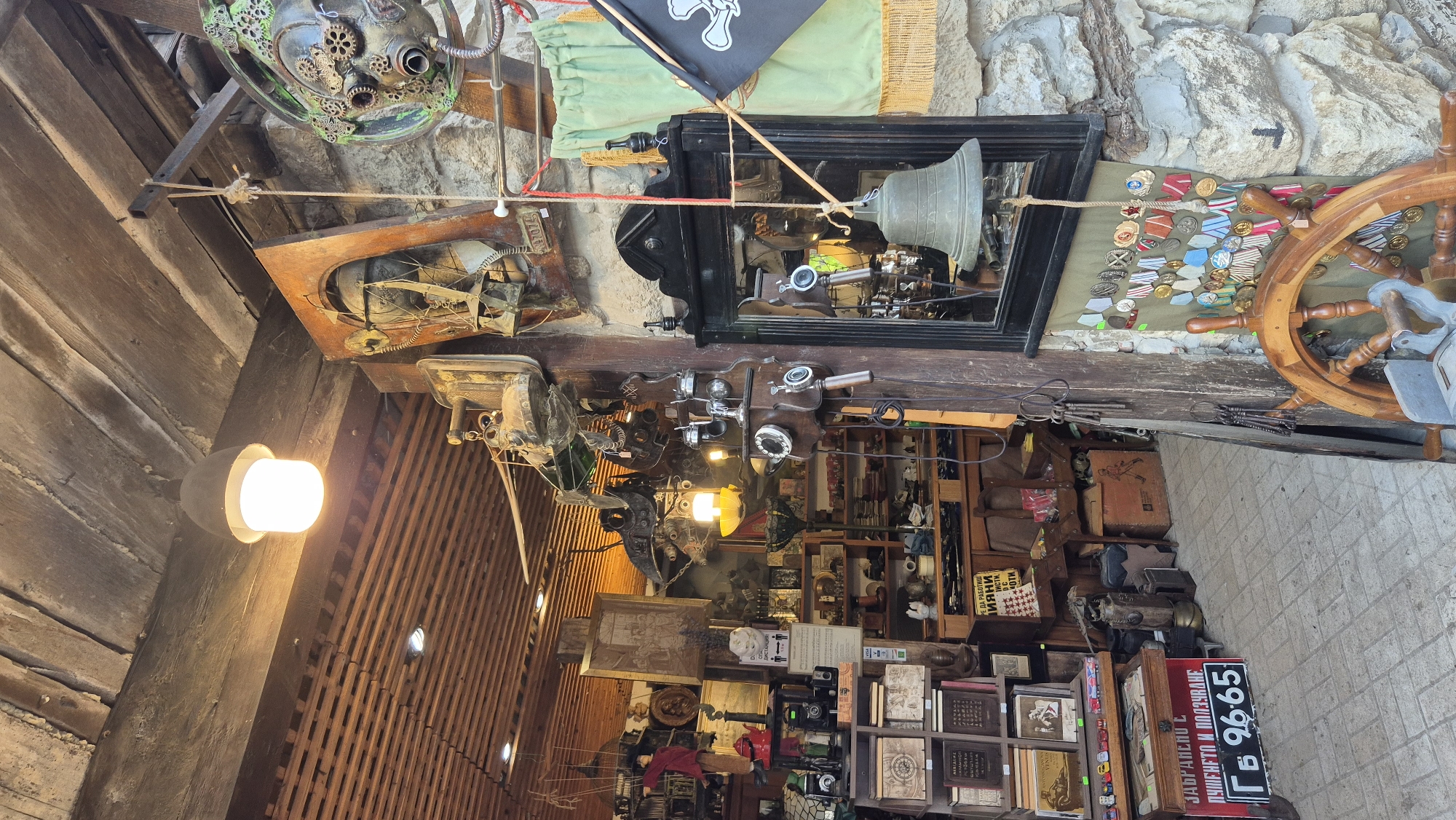The image depicts a dimly lit interior of a shop in the historic city of Nesebar, Bulgaria, likely an antique or souvenir store. The scene is cluttered with a variety of items, suggesting a collection of old artifacts and decorative pieces.
Dominating the upper portion of the image is a rustic wooden ceiling with exposed beams and wooden slats. A single, exposed light bulb hangs from the ceiling, providing a warm, artificial light.
The walls are a mix of rough-hewn stone and wooden paneling. Various objects are displayed on shelves and hanging from the walls and ceiling. These include:
* **Wall Decorations:** On the stone wall to the right, a green fabric hangs, adorned with a collection of medals. A black flag with a white skull and crossbones (pirate flag) is partially visible, attached to the right side of the green fabric. A large, dark-framed mirror is also present on this wall, reflecting some of the interior. Above the mirror, a bell is suspended by a rope. To the left of the mirror, a vintage-style wall-mounted telephone is visible.
* **Hanging Objects:** Suspended from the wooden beams and ceiling are numerous metal objects, many of which appear to be part of old machinery or artistic installations, with a steampunk aesthetic. There are also some mechanical-looking contraptions, possibly old tools or decorative elements. A puppet in a red garment hangs from wires on the far left.
* **Shelving and Displays:** Wooden shelves are packed with various items, including boxes, books, framed pictures, and smaller objects. Some boxes have decorative or informational labels. A prominent red sign with white lettering in Cyrillic alphabet is visible on the lower left, which appears to be a warning or informational notice.
* **Foreground Objects:** In the lower right foreground, a large wooden ship's wheel is prominently displayed. To the left of the ship's wheel, there are stacks of what appear to be books or albums.
The overall impression is of a densely packed, eclectic collection of items, creating a visually rich and somewhat overwhelming environment. The lighting is moody, contributing to an atmosphere of antiquity and mystery. No people are visible in the image.