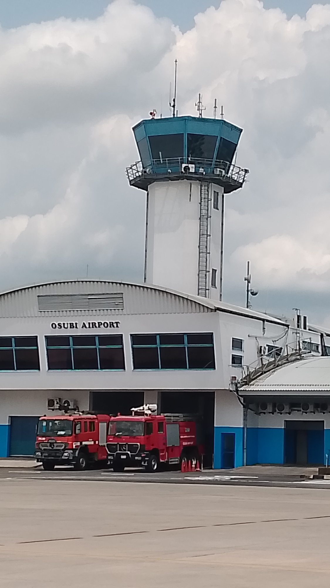 This media file captures a clear daytime scene of Osubi Airport in Orerokpe, Nigeria. The primary subjects are the airport's main building, its control tower, and two fire trucks.
The airport building, predominantly white, features a curved, corrugated metal roof. The name "OSUBI AIRPORT" is prominently displayed in black letters on the facade, above a series of large, dark-tinted windows. Below these windows, the building incorporates blue accents around what appear to be garage bays.
Parked within two of these bays are two bright red fire trucks, suggesting an active airport fire and rescue service. The truck on the left has "MAN" visible on its grille, indicating its manufacturer. The truck on the right has faint text, "RESCUE SUPPORT," visible on its side.
To the right and slightly behind the main building stands a tall, white control tower. Its cylindrical shaft is equipped with an external ladder. The control cab at the top is octagonal, featuring numerous windows framed in a distinct teal-blue color. Various antennas and communication devices are mounted on and around the control cab.
The sky is partly cloudy, with large, white cumulus clouds visible, indicating fair weather conditions during the day. The scene depicts a functional airport facility, with the fire trucks ready for deployment, underscoring the operational nature of the location.