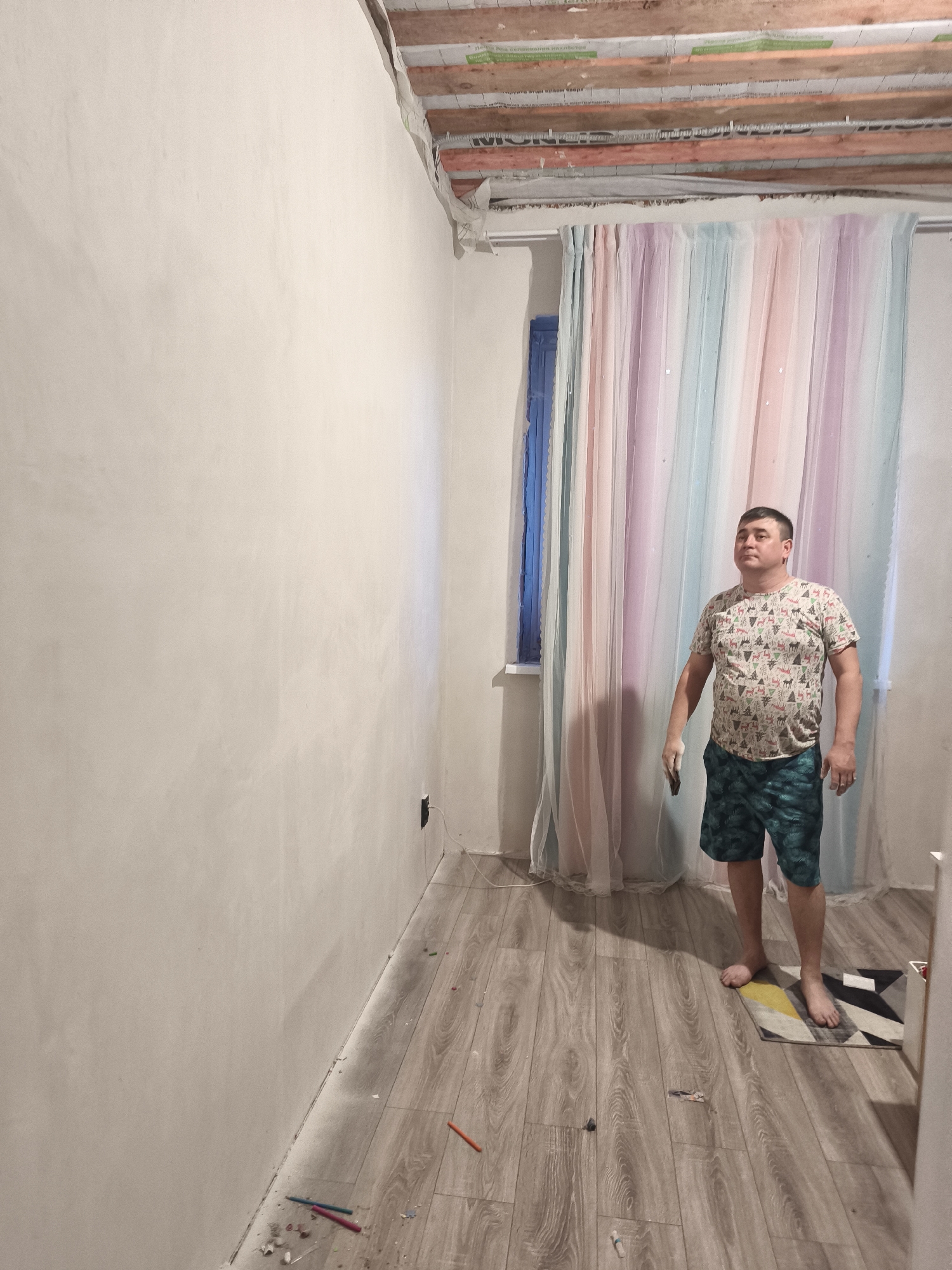 The photograph depicts a room under renovation, likely located in Сокуры, Russia, given the information provided. The left wall is freshly plastered and painted a light color. The ceiling is unfinished, exposing the wooden beams and insulation materials. A window is visible on the right side of the room, covered by multi-colored sheer curtains. A man stands near the window, holding a small object in his hand. He is wearing a patterned t-shirt and green shorts. The floor is covered with wood-patterned flooring, with scattered debris and tools visible. A small rug is partially visible in the lower right corner. The overall impression is that of an ongoing home renovation project.