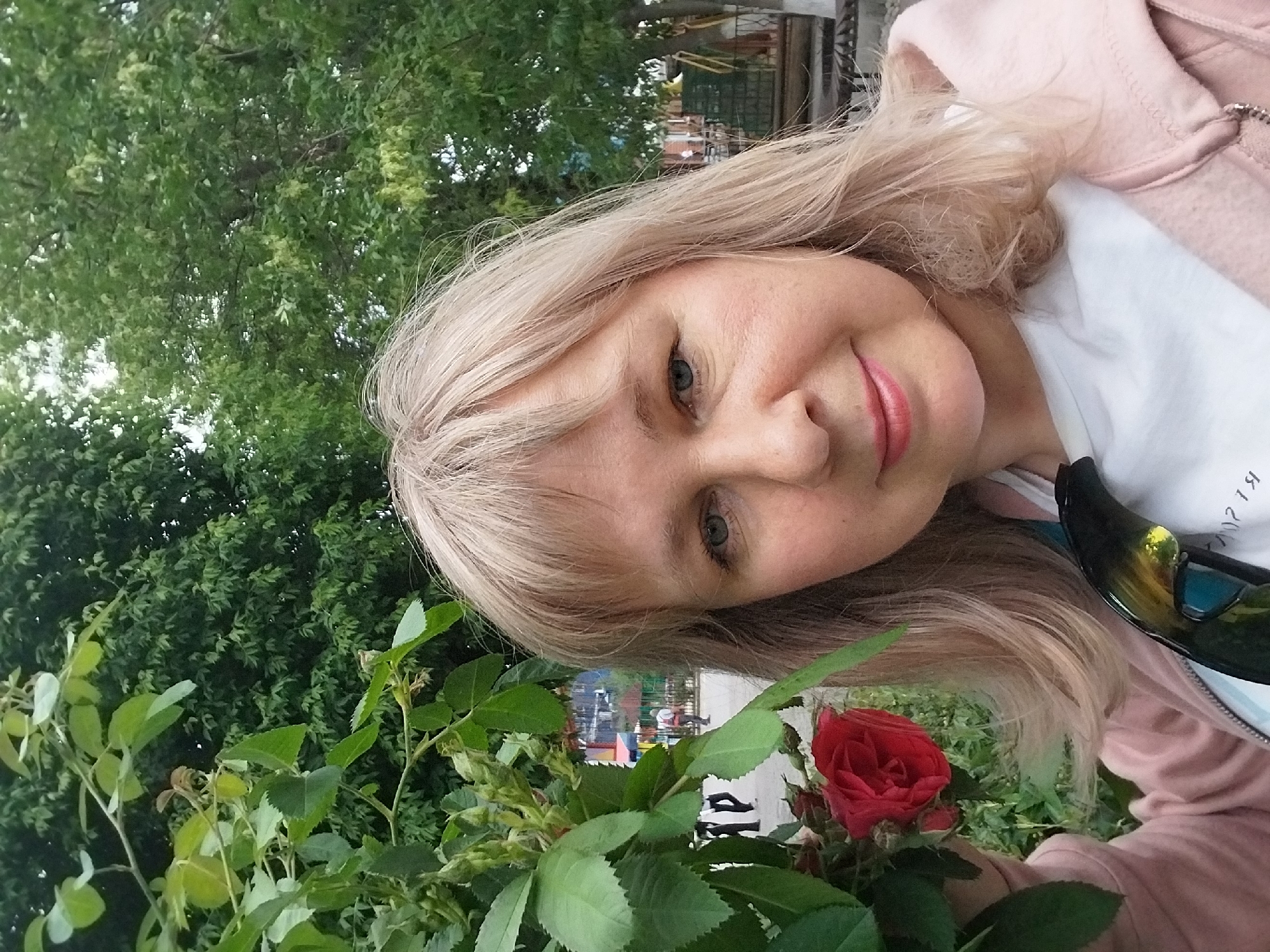 The image shows a woman with blonde hair, wearing a pink hooded jacket over a white shirt, posing outdoors with a red rose. The background features lush green foliage, suggesting a park or garden setting. In the blurred background, there are glimpses of urban elements like a playground, indicating the photo was likely taken in a city park. The location is Orenburg, Russia.