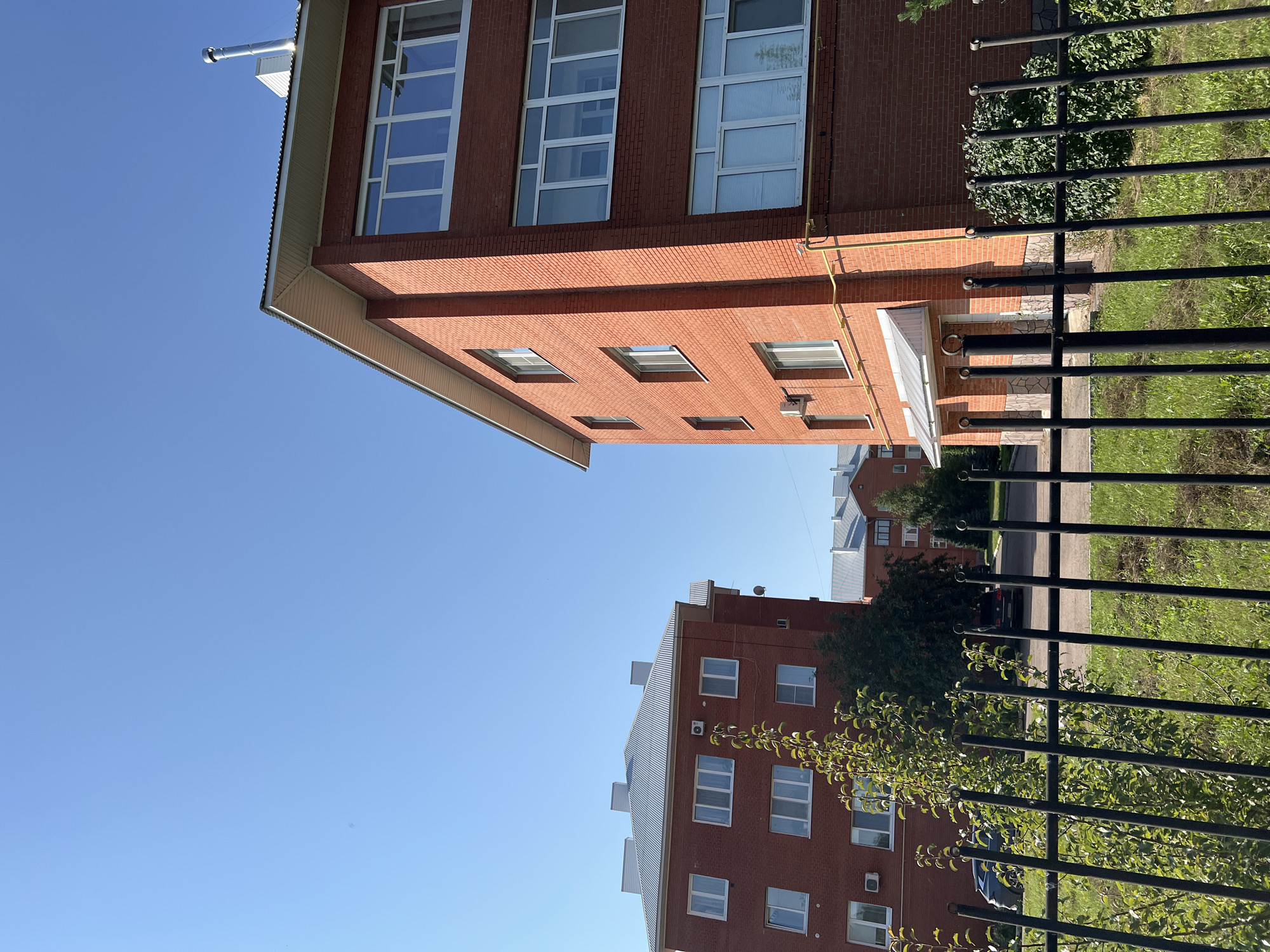 A low-angle, eye-level shot captures a clear, bright blue sky over a residential area in Salavat, Russia. Two red brick apartment buildings dominate the frame, with the building on the right in the foreground. This building features multiple floors with white-framed windows, some of which have blinds. A silver chimney stack rises from the roof of this building. The building on the left, also brick, is partially visible and appears to have a grey, corrugated roof. Between the buildings, a few trees and green bushes are present, along with a black metal fence in the foreground. The ground appears to be covered in grass and a paved area, suggesting a walkway or road. Two dark-colored cars are parked in the background on the paved area. The scene is well-lit, indicating daylight.