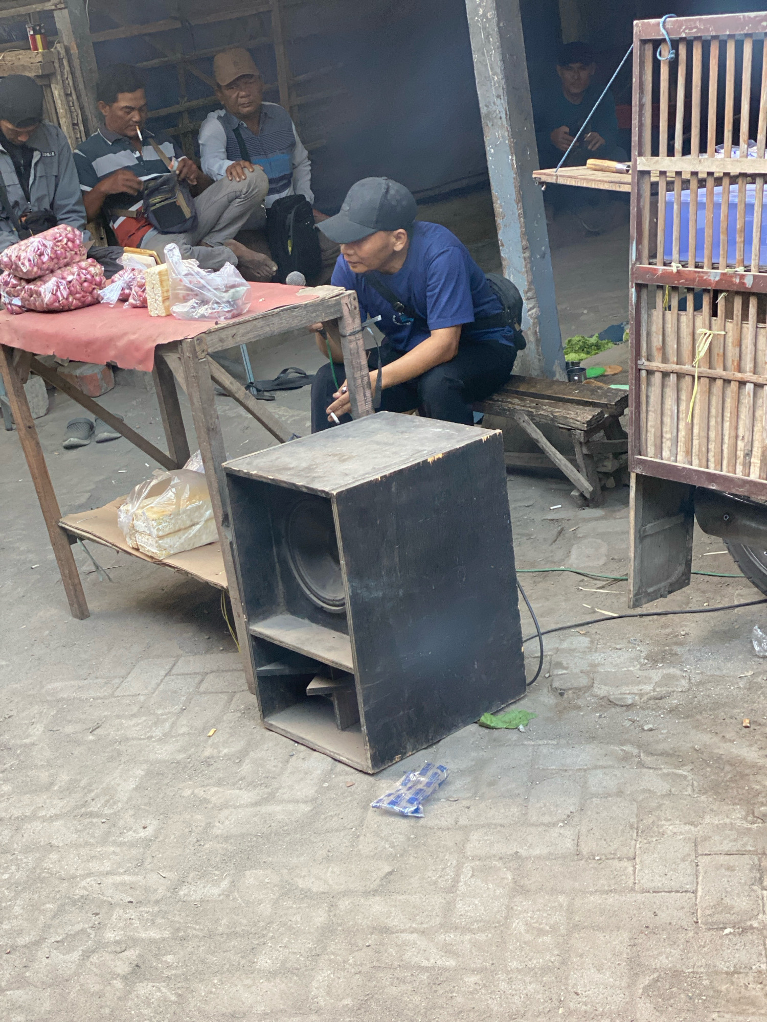 In Gurah, Indonesia, a group of men are gathered outdoors, under a makeshift shelter constructed of rough wood. A table covered with a red cloth displays several bags of red onions and some packaged food items. One man, seated and wearing a blue t-shirt and a dark baseball cap, is looking down, holding something in his hand. To his left, another man in a striped shirt and cap smokes a cigarette. Further back, two other men are visible. In the foreground, a large black speaker sits on the ground. The environment appears to be a market or roadside stall, with a paved ground surface and weathered wooden structures.
