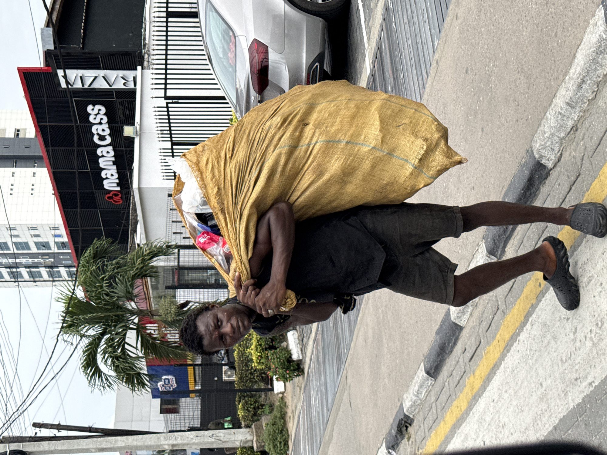A man carries a large yellow bag on his back, walking along a sidewalk. The sidewalk borders a street and a building with the sign "mama cass" on the side. The location appears to be in Itirin, Nigeria, based on the visible sign.