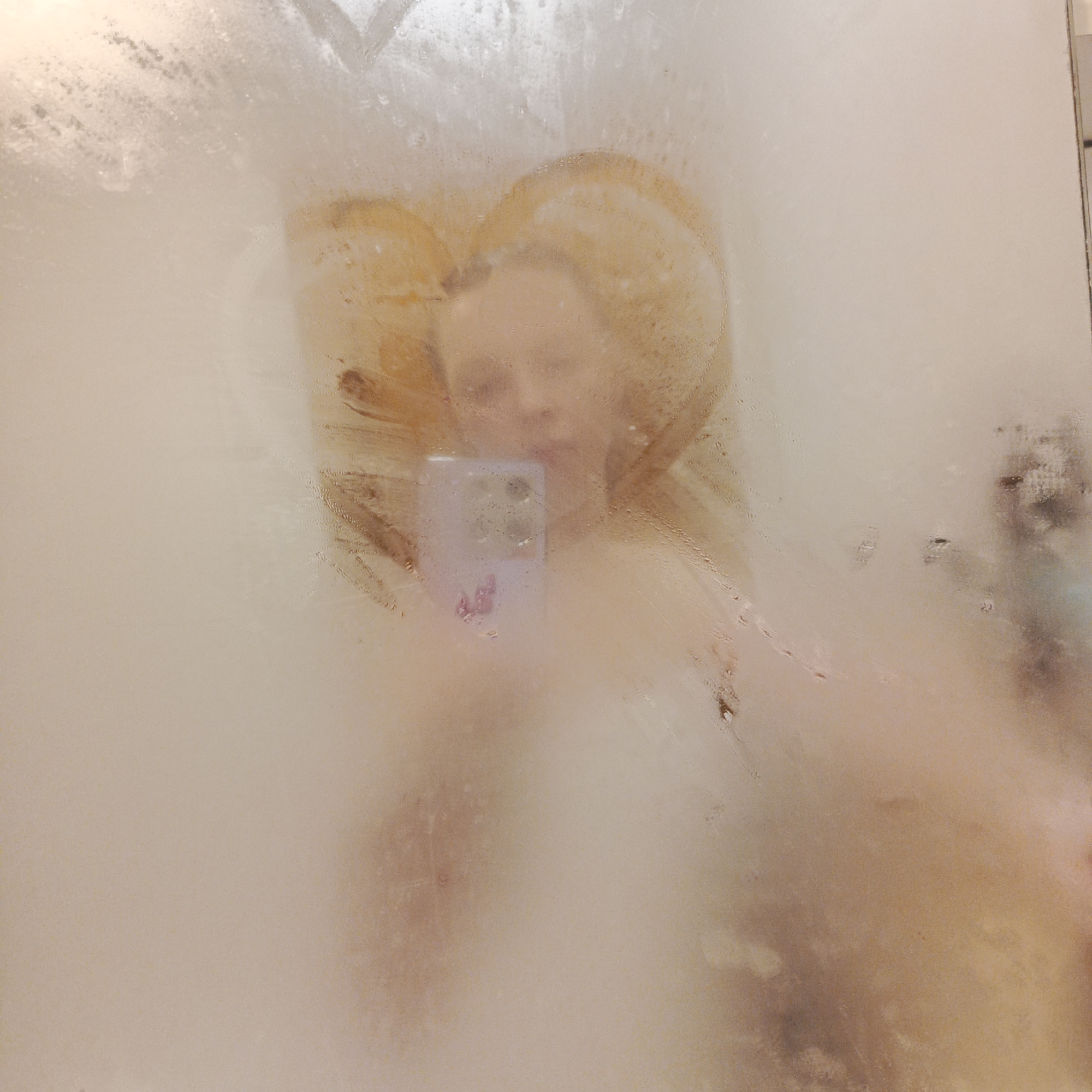 A woman is taking a selfie in a steamy shower. Her blonde hair is visible through the condensation, and she is holding a phone with a purple case. The background is a blurred, tiled shower wall. No information regarding the location in Chaikovsky, Russia is available.