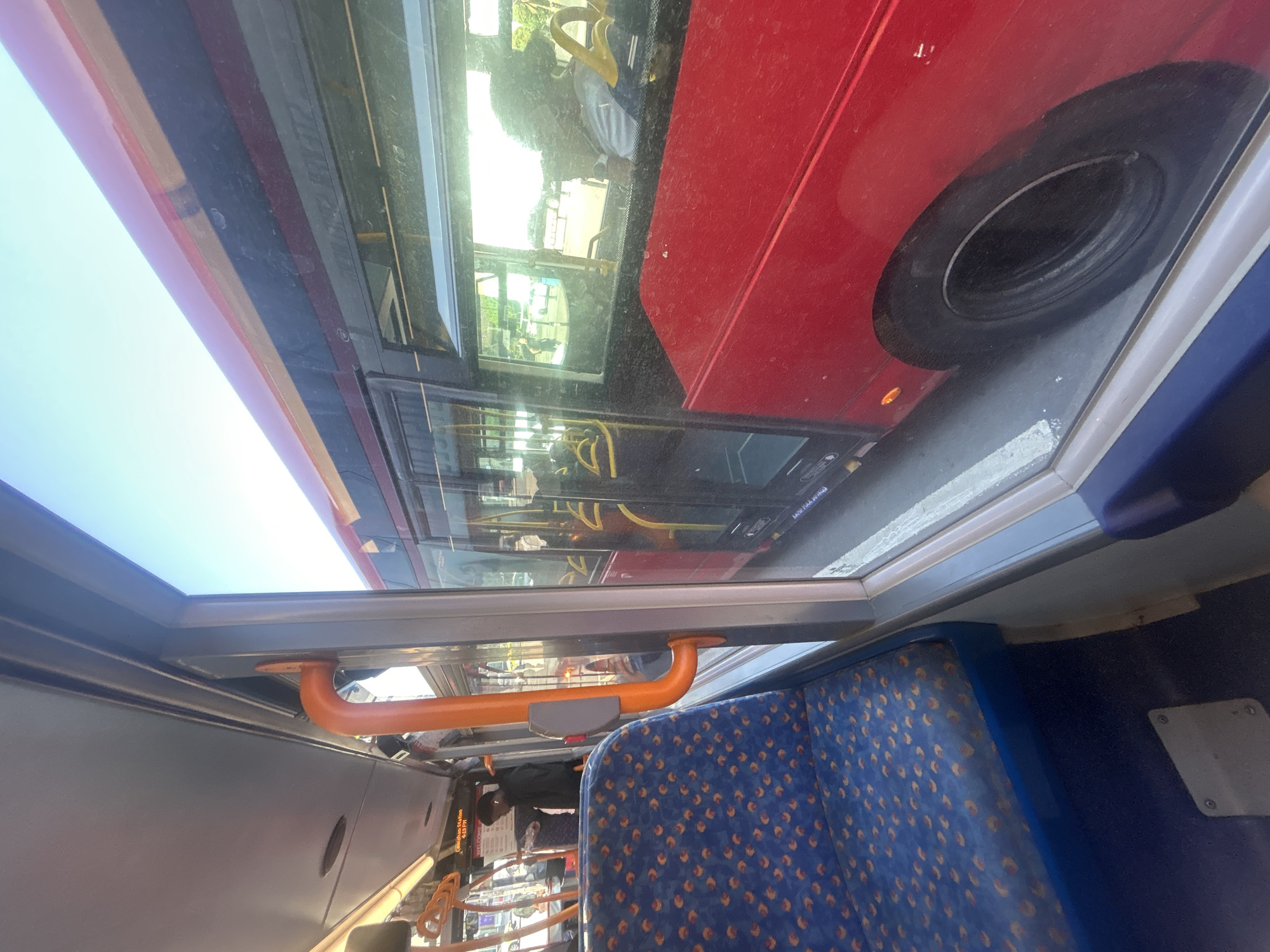 The image, taken from the upper deck of a bus in London, UK, shows the interior of the bus and a view out of a window. Inside, there is blue patterned seating and orange handrails, indicative of London public transport. The window view reveals another red London bus alongside, with its black tire visible. The reflection in the window contains the scene outside. The overall location is confirmed by the style of the bus and familiar interior design.