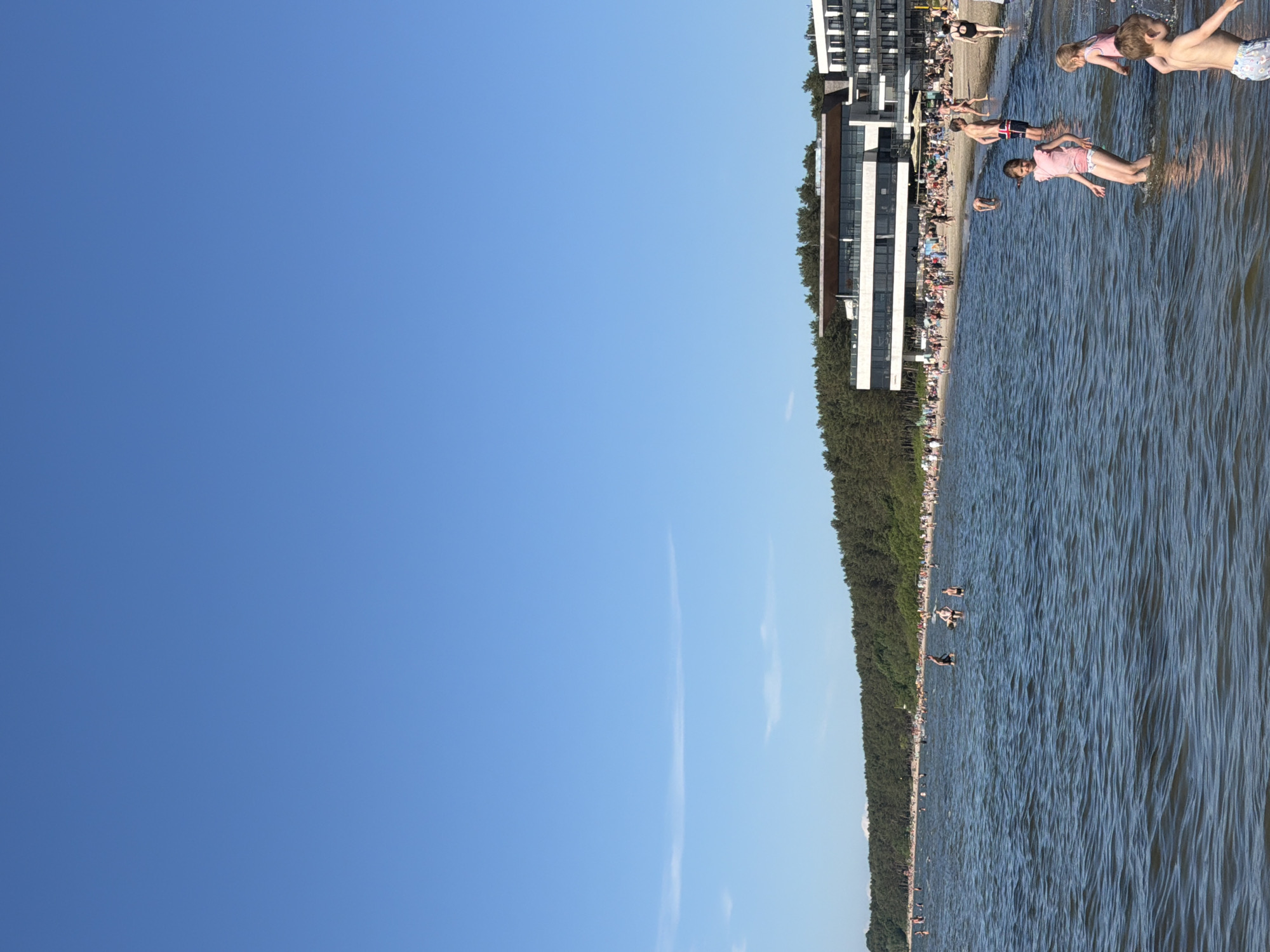 A vertical, eye-level medium shot captures a crowded beach on a clear day. In the foreground, several children are wading in shallow, wavy water. To the right, a young boy with light brown hair wears patterned shorts and is partially submerged in the water, facing right. Next to him, a young girl with blonde hair wears a pink swimsuit and stands facing left, her arms outstretched. Further to the left, another young girl with blonde hair and a pink swimsuit stands with her arms by her sides, facing left. In the mid-ground, the sandy beach is densely populated with people relaxing and sunbathing. Behind the beach, a dense line of green trees forms a backdrop. On the far right of the mid-ground, a modern, multi-story white building with dark windows is visible. In the far distance, a clear, bright blue sky fills the upper portion of the image. The scene is consistent with a popular beach location in Tallinn, Estonia, possibly Pirita Beach.