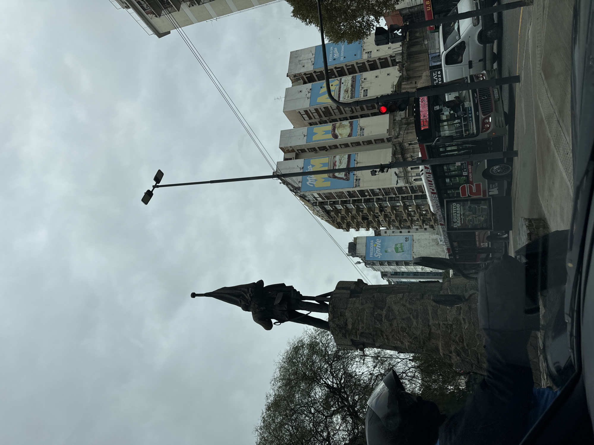 The image shows a view from inside a vehicle, likely a car, looking out onto a city street in Buenos Aires, Argentina. The focal point is a monument featuring a statue of a man holding a flag, set upon a stone base. Trees surround the base of the monument. Above the monument, the sky is overcast.
In the background, tall buildings flank the street. Large advertisements for "Milka" and "Sprite" are visible on the sides of the buildings. A traffic light, showing a red light, is present. A public bus is partially visible, displaying information about "Transportes Automotores Callao S.A." and the bus number "61" and "BA". A white van is parked further down the street.
The vehicle's interior and side mirror are partially visible in the foreground, adding to the perspective. The overall scene captures an urban landscape on what appears to be an overcast day.
