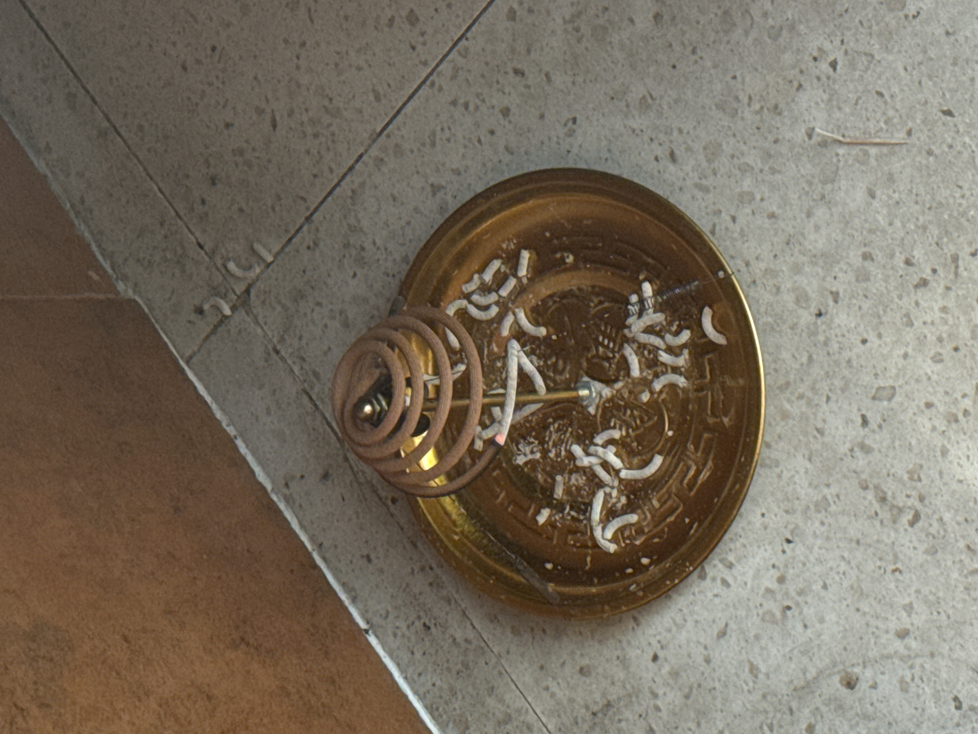 The scene is indoors, possibly in a business, and located in North Kuta, Indonesia. The foreground shows a golden incense burner on a tiled floor. The burner contains the remains of burnt incense coils, primarily white ash remnants. A burning incense coil is positioned on a metal stand above the ash. The surrounding floor is tiled with mottled grey-brown tiles, and the bottom-left corner of the image reveals a different, smoother brown flooring material.