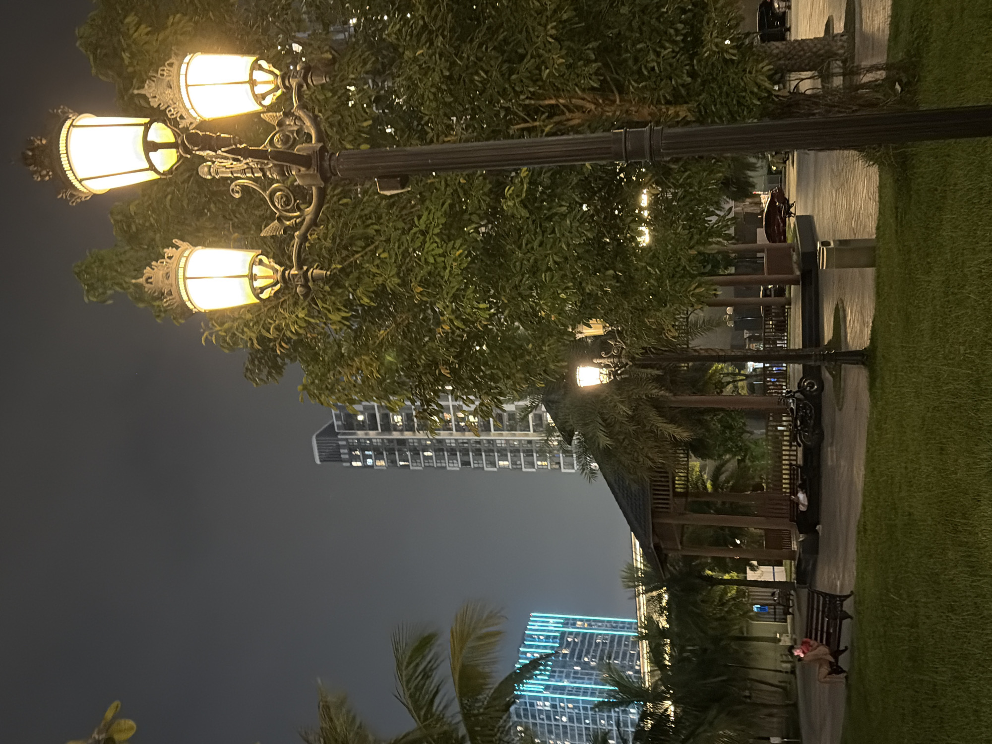 This is a nighttime scene in a park, likely in Hanoi, Vietnam, given the architectural style of the buildings in the background which appear to be modern high-rises. A large, ornate lamppost with three illuminated lamps stands prominently in the foreground, casting a warm glow on the surrounding trees and pathway. To the left, a gazebo structure is visible, with palm trees and other lush greenery interspersed throughout the park. Several people are present in the scene: one person is sitting on a bench in the mid-ground, another is lying down on a bench near the gazebo, and a motorcycle is parked nearby. In the background, several tall, modern buildings are illuminated, some with distinct blue lighting on their facades, suggesting a commercial or residential district. The overall ambiance is serene and urban, with the artificial lighting creating a distinct atmosphere.