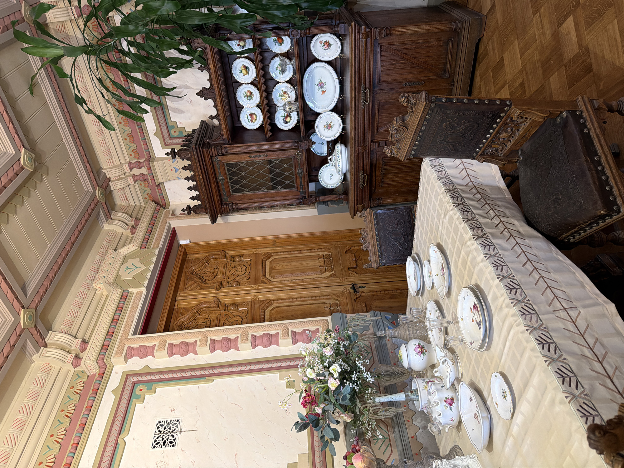 This image depicts an opulent dining room, likely within a historic building in Nizhny Novgorod, Russia. The scene is set with a table laid for a meal, showcasing a collection of fine china, crystal glassware, and a floral centerpiece. A large, ornate wooden hutch stands against the wall, displaying a variety of decorative plates, some with fruit motifs and others with floral designs. The room is richly decorated with intricate ceiling moldings, elaborate wall ornamentation, and richly carved wooden doors. The furniture, including a dark wood hutch and antique chairs upholstered in leather, suggests a bygone era. A large houseplant in the corner adds a touch of greenery to the scene. The overall impression is one of grandeur and historical significance.