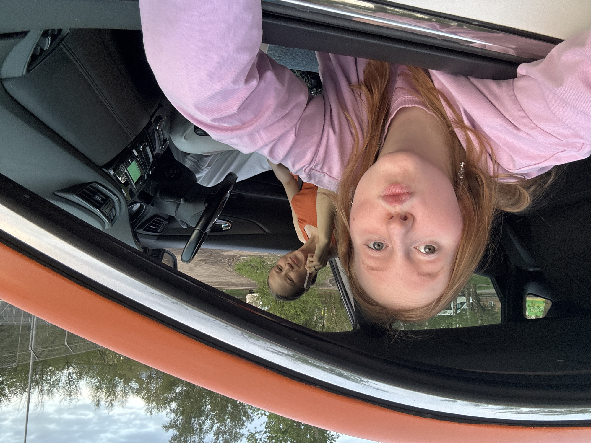 Two women are inside a car, likely parked. One is in the driver's seat and the other is leaning out of the window, looking at the camera. The car appears to be orange. The background through the windows shows a cloudy sky and some trees. The environment appears to be located near a fence and some structures, possibly buildings, in an urban or semi-urban setting. No verifiable information indicates the location is Perm, Russia.