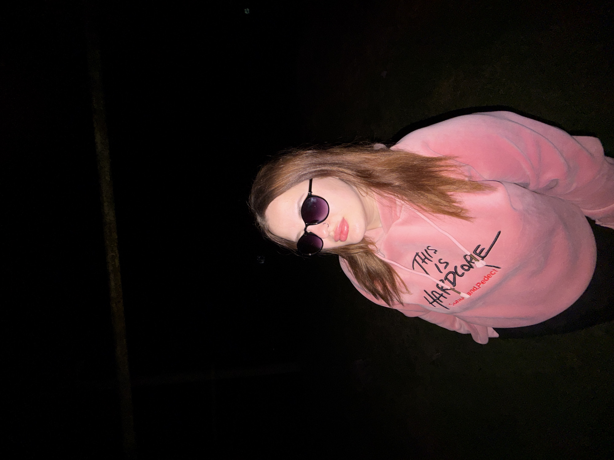 A woman with brown hair and sunglasses is wearing a pink sweatshirt with the words "THIS IS HARDCORE" on it. She is standing outdoors at night. The location cannot be determined as being within the city of Novopokrovskaya, Russia, due to the image lacking contextual information.
