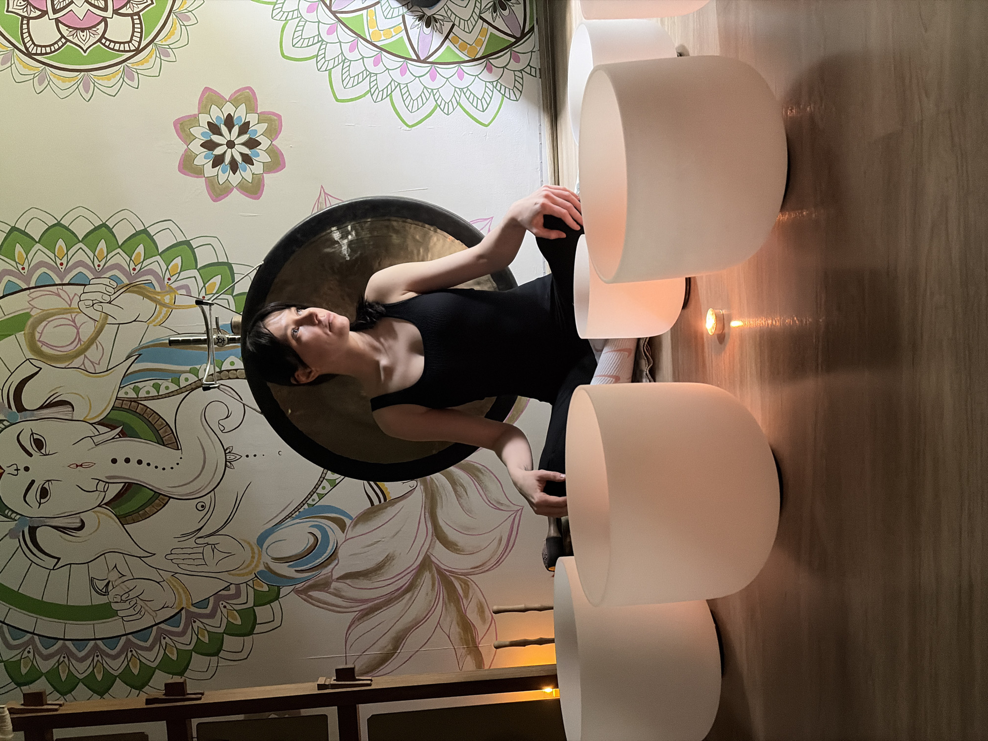 An indoor scene in Chaikovsky, Russia, features a female individual seated on a light brown wooden floor. The woman, with dark hair, wears a black ribbed tank top and black leggings with white socks, positioned in a cross-legged posture. Her hands rest on her knees, palms down, and her head is turned to the left, directing her gaze out of frame. Directly behind her is a large, dark, circular gong mounted on a metal stand. In the foreground, five frosted translucent crystal singing bowls of varying sizes are arranged in a semicircle on the floor, emitting a warm glow. A small, lit tea light candle is visible on the floor between the bowls, casting reflections on the wooden surface. The white wall behind the woman is adorned with a colorful mural, prominently featuring a large depiction of Ganesha, a Hindu deity with an elephant head and multiple arms, on the left. The mural also includes various circular mandala patterns and lotus designs in shades of green, pink, yellow, blue, and brown. A small mallet or striker is present near the woman's right leg. Dark wooden structural elements are partially visible on the far left side of the frame.