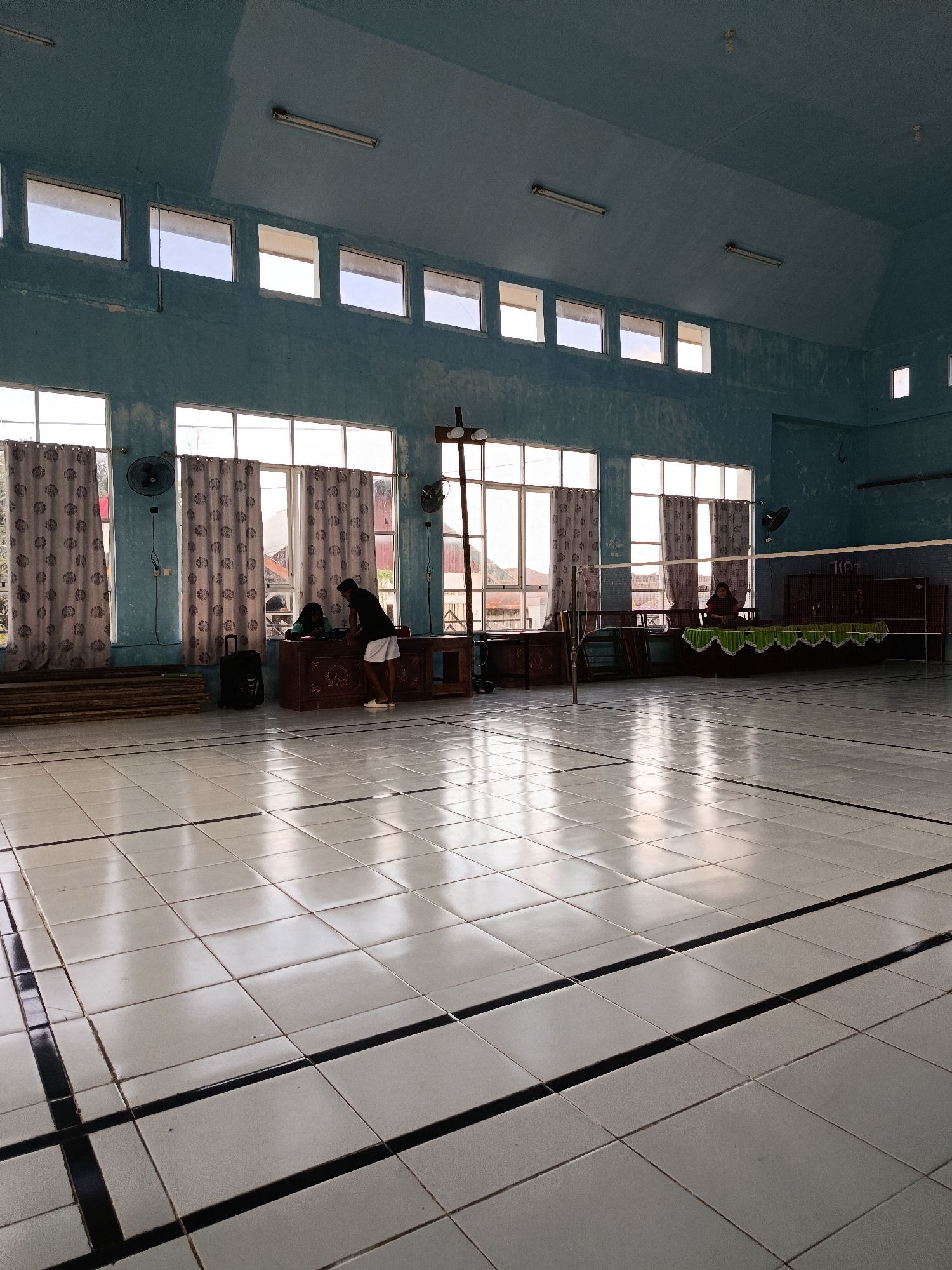 This is an indoor sports facility, possibly a badminton court, located in the city of Mansalean, Indonesia. The room has a light blue ceiling with fluorescent lights. Large windows with patterned curtains line the upper portion of the walls, allowing natural light to enter. The floor is tiled with white square tiles, marked with black lines to delineate courts. In the foreground, the tiled floor reflects the light from the windows and ceiling.
In the mid-ground, several objects and people are visible. There are wooden benches and desks along the walls. A black suitcase stands near a desk where two individuals are interacting. One person, wearing a black shirt and white shorts, is leaning over the desk. Another person, mostly obscured, appears to be seated behind the desk. Further back, a badminton net is set up across the court. On the far side of the court, another individual is seated behind a long, ornate wooden structure, also draped with a green cloth. Two black electric fans are mounted on the walls, one on the left and one on the right. Wooden planks are stacked along the left wall. The overall environment suggests a functional space for sports and potentially community gatherings.