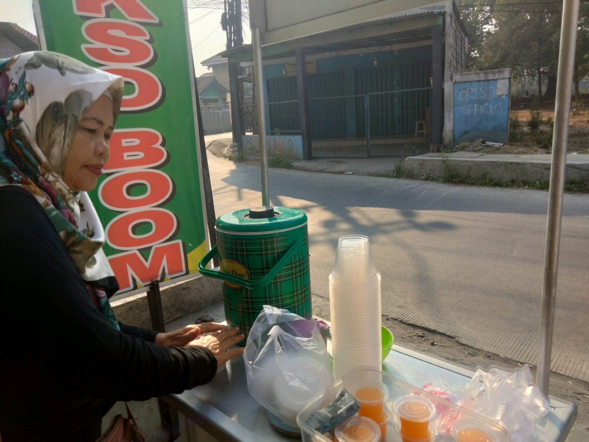 In the city of Pisangan Jaya, Indonesia, a woman wearing a headscarf stands behind a street food stall. She is in the process of serving drinks. In front of her, a green plaid insulated cooler is visible, along with a stack of clear plastic cups and several small plastic cups filled with orange liquid, likely juice. There is also a plastic bag filled with ice. In the background, a blue building with metal gates suggests a commercial area. The street is paved and appears to be a public thoroughfare. The lighting indicates it is daytime, possibly late afternoon, with long shadows cast by the sun.