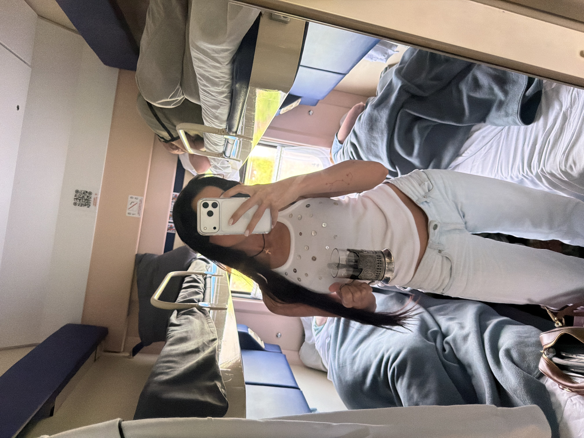 A woman is taking a mirror selfie in a train compartment. She is holding a phone in her right hand and a glass with a metal holder in her left hand. She is wearing a white tank top with sequins and light-colored pants. The train compartment has bunk beds with blue and dark gray bedding. Another person is visible lying on an upper bunk in the background. The scene appears to be set within a train traveling through a rural or suburban landscape, indicated by greenery visible through the window. The location is identified as Babyakovo, Russia.