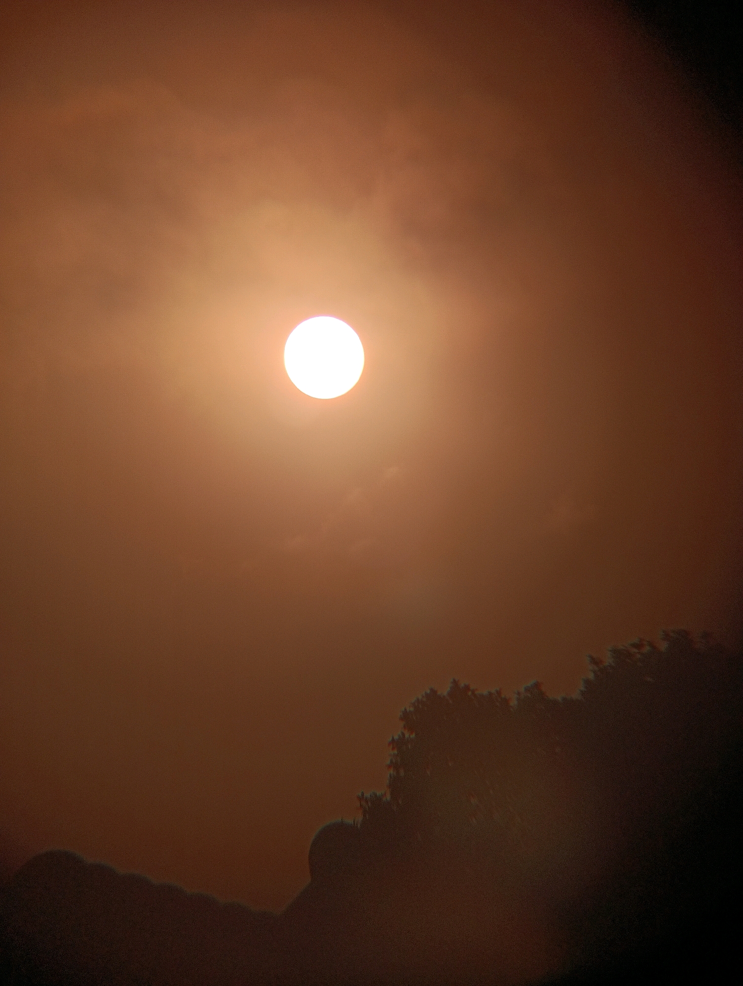 A bright, white sun is visible in a hazy, orange sky. In the foreground, silhouetted against the sky, are the tops of trees and what appear to be hills or buildings. The image was taken in Pematang Jaya, Indonesia.