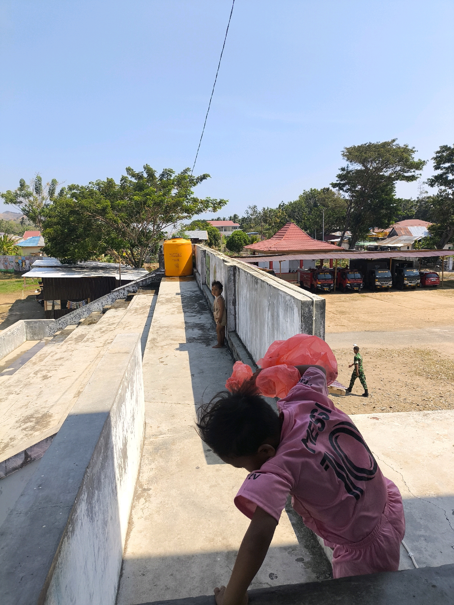 In Tilamuta, Indonesia, on a bright, sunny day, a young child, seen from behind and wearing a pink shirt with "MESSI 10" printed on the back, leans forward on a concrete surface. The child is holding two bright red plastic bags. To the left, another child, dressed in light brown shorts and a short-sleeved shirt, stands near a tall, bright yellow water tank situated on a flat concrete area. Behind the children and the water tank, a long, low-lying building with a corrugated metal roof is visible, and beyond that, a cluster of houses with various colored roofs. In the distance, a line of parked red fire trucks is under a covered structure. Further in the background, trees and a gentle hillside can be observed. A person in a green military uniform walks away from the fire trucks. The overall scene depicts an outdoor area with a mix of residential buildings, service infrastructure, and natural elements.