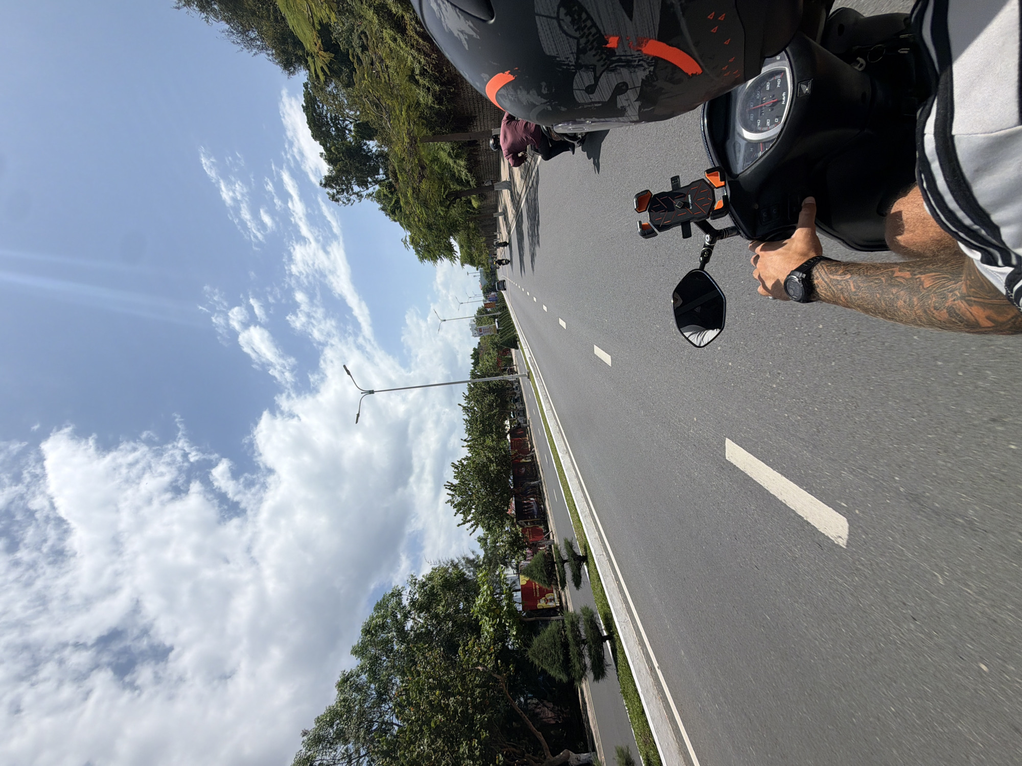 The image presents a first-person perspective from someone riding a scooter on a clear, sunny day in Bac Nha Trang Ward, Vietnam. The foreground shows the rider's right arm, which is heavily tattooed, holding the handlebar of a black scooter. A black digital watch is visible on their wrist. The rider is wearing a black helmet with orange and grey patterns, and a white and grey striped short-sleeve shirt.
The scooter's dashboard, displaying a speedometer marked with "km/h" and other indicators, is prominent. A "ROCK BROS" brand phone holder is attached to the handlebar. The road ahead is a wide, paved asphalt stretch with multiple lanes, clearly marked by white dashed lines. Several other scooters or motorbikes can be seen in the distance, also traveling along the road.
The sides of the road are lined with green verges and numerous trees with dense foliage, suggesting a tropical or subtropical climate. Beyond the trees, some indistinct buildings and billboards with unreadable text are visible. The sky is bright blue with scattered white cumulus clouds, indicating fair weather. The sun is high, casting distinct shadows on the road and from the trees. The overall scene conveys a calm journey on a well-maintained road under pleasant conditions.