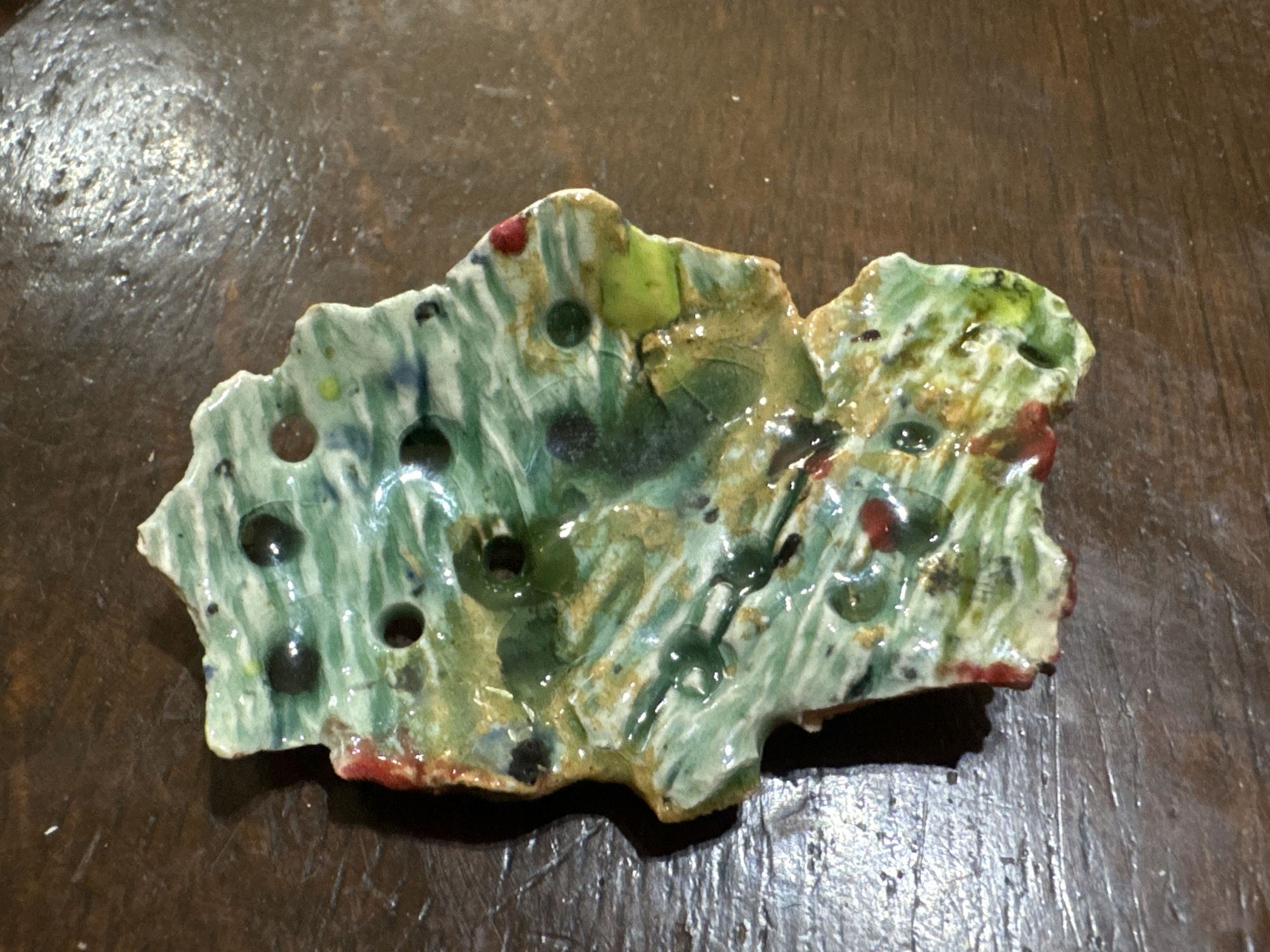 An abstract ceramic shard, irregularly shaped and primarily green with streaks of white and yellow, lies on a dark, textured wooden surface. The shard features several small, circular indentations, some filled with dark glaze. There are also scattered red and dark blue accents within the glaze. The lighting casts a soft shadow to the left of the shard. The location is not specified and therefore is Unknown, Unknown.