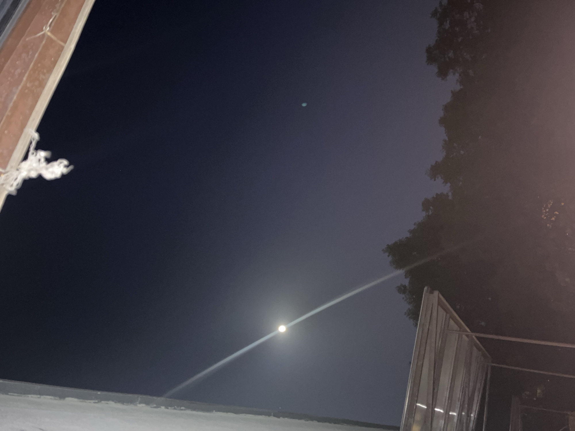 The image depicts a night scene with a clear sky, possibly in a city. A bright moon is visible in the lower center of the frame, with a faint light streak extending horizontally across the sky. A building or structure is partially visible on the left side, while a tree with dense foliage is on the upper right. A metallic or reflective structure is on the right-lower area of the frame. The location is Unknown, Unknown.