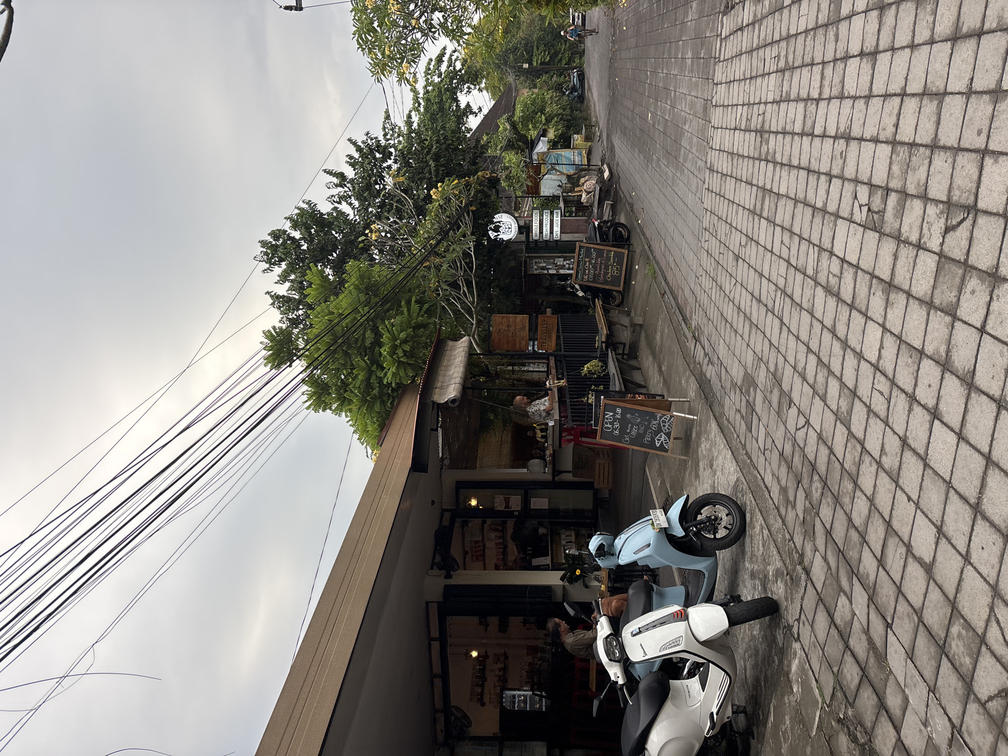 In Ubud District, Indonesia, a street scene unfolds under an overcast sky. Two scooters, one white and one light blue, are parked on the textured, grey cobblestone street in front of a building housing a cafe and market. The building has a light brown, sloped roof and large glass windows, through which interior shelves stocked with items are visible.
Inside the cafe, two individuals are seated, one man looking down at his phone and another man interacting with a staff member behind a counter. A blackboard sign outside the cafe advertises "OPEN 06:30 - 16:00", "Get some Coffee and Pastry 60K".
Further down the street, more signs are visible, including one that reads "COMMUNITY MARKET" with a logo, and another with text indicating "WHOLE FOODS", "FRESH PRODUCE", and "CAFE - JUICE BAR". A second, smaller blackboard displays prices in what appears to be Rupiah. Another person is seen cycling away from the camera down the street. Lush green trees and foliage overhang the buildings and street, contributing to the tropical atmosphere. Electrical wires are strung across the upper portion of the frame. The overall environment suggests a relaxed, open-air commercial area in a tropical climate.