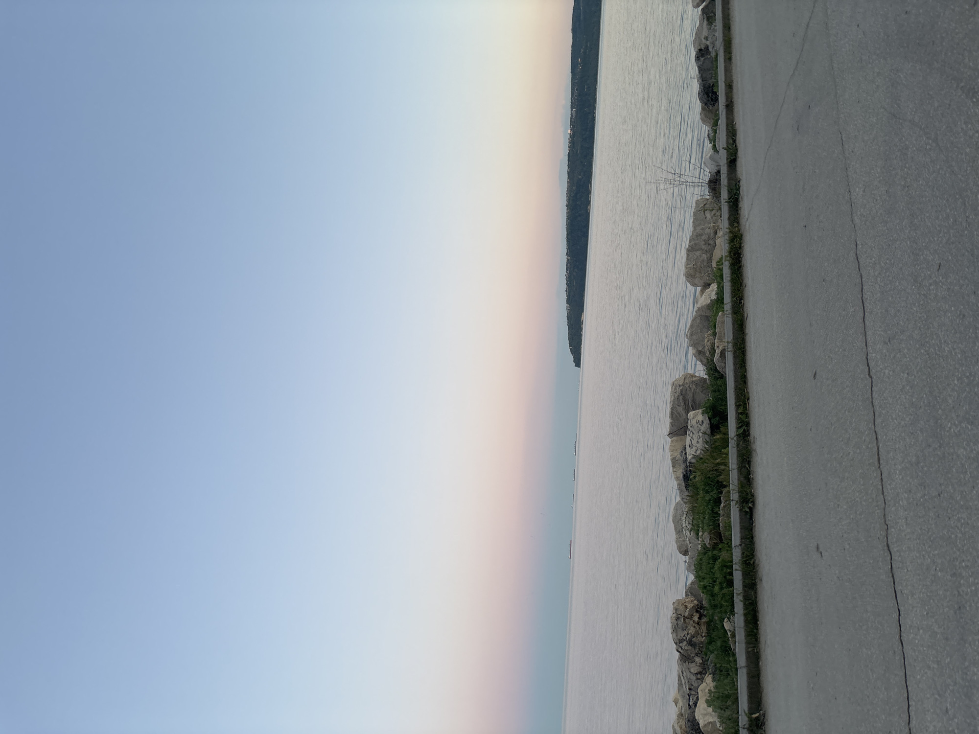 The image depicts a coastal scene in Varna, Bulgaria, at dusk. On the left, the sky transitions from light blue to a pale orange-pink horizon. The water of the Black Sea occupies the central part of the image, with a tree-covered coastline visible in the distance. The right side of the frame shows a paved road, separated from the water by a row of large rocks and some green vegetation. The road has a visible crack running along its surface. The scene is devoid of any visible people or significant activity. The overall mood is tranquil.