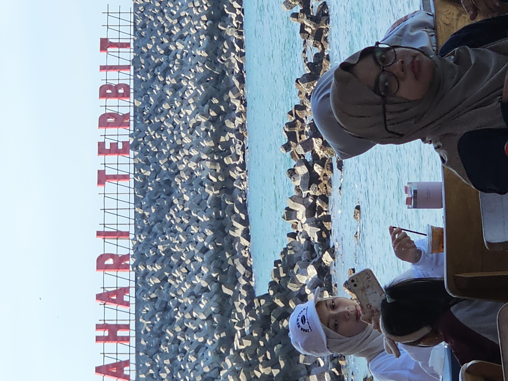 This image shows a group of people near a beach in Denpasar, Indonesia. In the background, a sign with large red letters spelling "AHA RITTERBIT" is visible, likely referring to the Sunrise Beach or a similar location. The beach is lined with tetrapods, concrete structures designed to prevent coastal erosion.
In the foreground, two women are seated at tables. The woman on the left is wearing a white top and a light-colored hijab and a cap. She is holding up a smartphone, possibly taking a picture or video. In front of her is a cup with a straw, and a pink water bottle. To her right, a younger person with dark hair is visible. The woman on the right is wearing a brown hijab and glasses. She is looking towards the camera. Other people are partially visible in the background. The water is a clear blue, and the sky is bright.