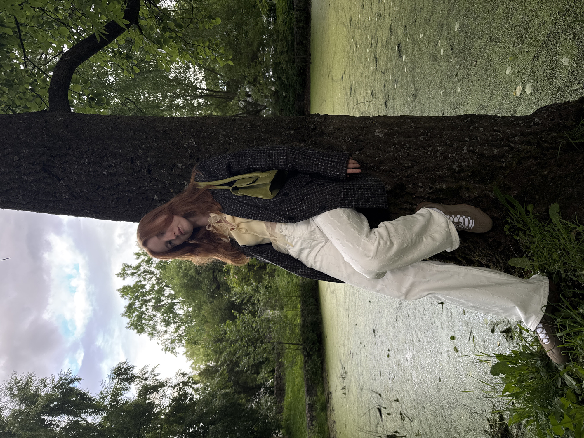 A young woman with red hair is leaning against a large tree trunk. She is wearing a light yellow blouse with a tie detail at the neck, a dark plaid blazer, and loose white patterned trousers. She has a small, lime green shoulder bag. Her left leg is bent, and her right foot is resting on the ground. The tree is positioned next to a body of water covered in green algae, with some lily pads visible. In the background, there are trees and a cloudy sky. This scene appears to be in a park or garden setting.