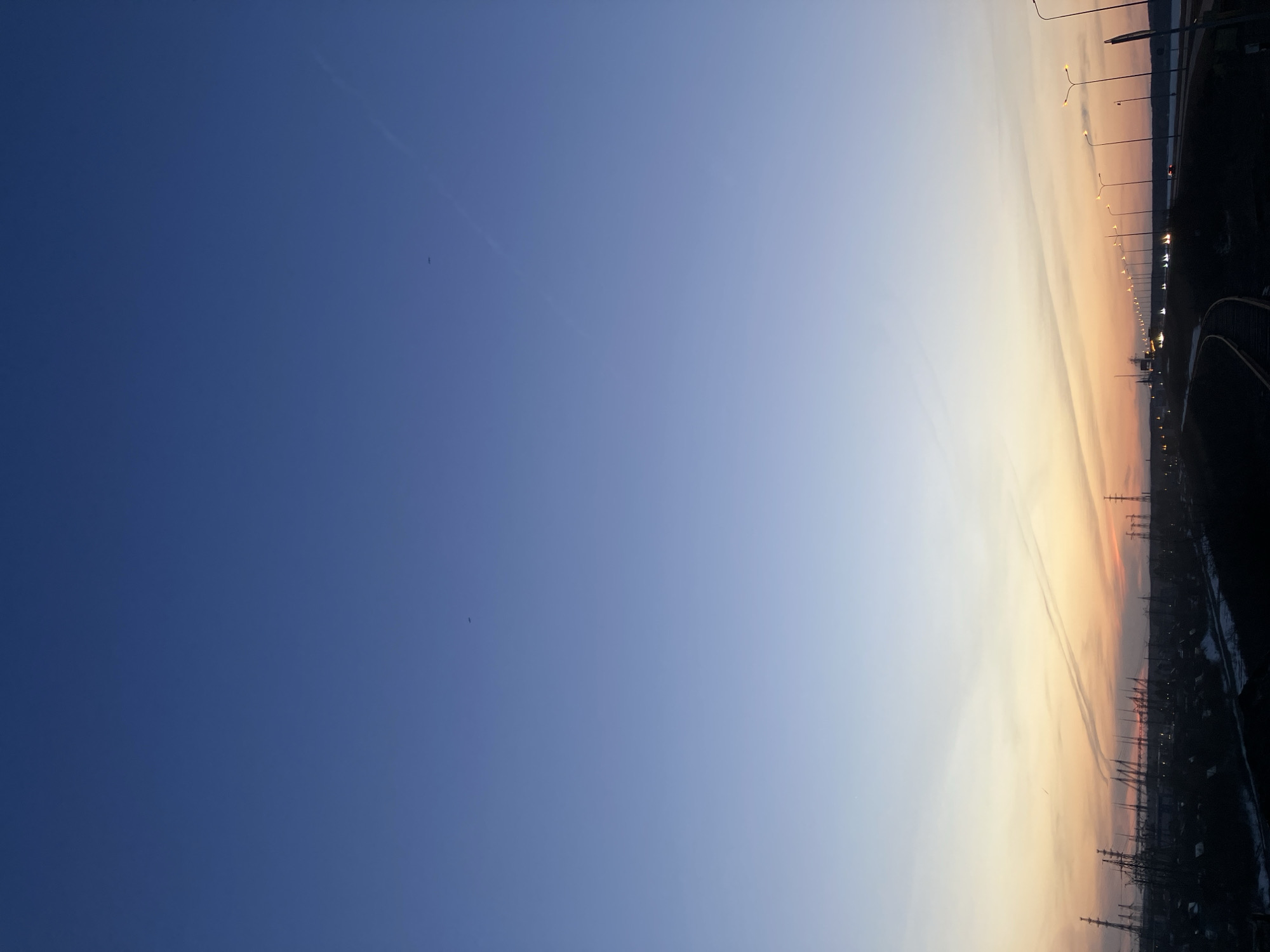 The image shows a view from a high vantage point, likely in Chaikovsky, Russia, due to the presence of infrastructure consistent with that location. The sky transitions from deep blue to lighter shades of blue and orange as it descends toward the horizon. On the right side of the image, a silhouette of a city skyline is visible, including power lines, buildings, and streetlights, suggesting an urban or industrial area. A road or bridge is also visible on the right.