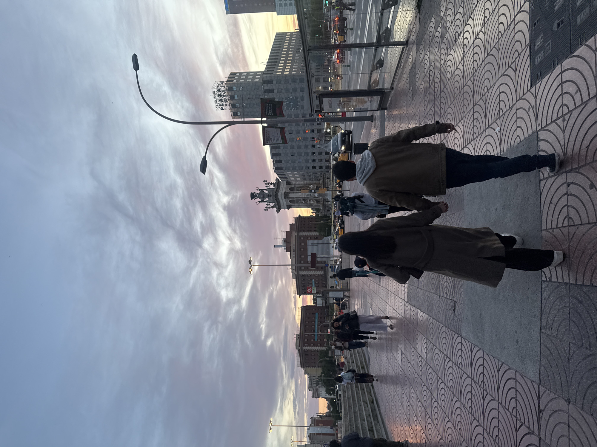 Here is a description of the image:
The image shows a street scene in Barcelona, Spain, likely at Plaça de Catalunya. Two people are walking on a patterned sidewalk, a woman in a long coat and a man in a jacket. Behind them, other people are walking along the pedestrian area. In the background are buildings, including a building with a clock tower that says “Catalonia” on it. A monument with statues can be seen in front of that building. Streetlights are visible along the street. The sky is a cloudy, pastel color, indicating that it is either dawn or dusk. Cars and buses are visible on the street.