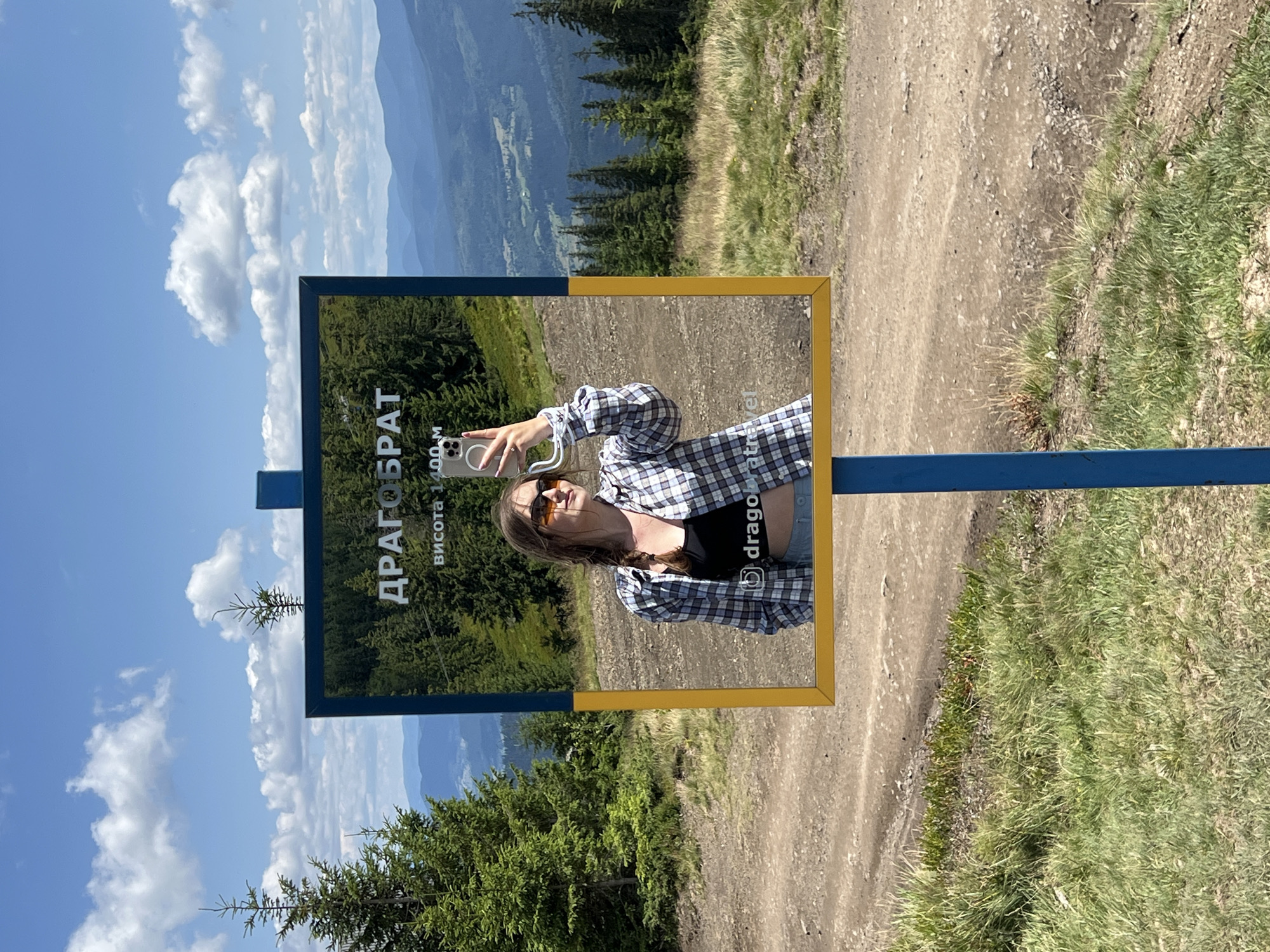 In Chorna Tysa, Ukraine, a young woman with braided brown hair, wearing sunglasses, a plaid shirt over a black top, and denim shorts, takes a selfie in a reflective sign. The sign displays the Cyrillic text "ДРАГОБРАТ" and "висота 1400 м," indicating "Dragobrat" and "height 1400 m." The sign has a blue pole and a yellow frame. The background reveals a mountainous landscape with lush green pine trees, rolling hills, and a clear blue sky with white clouds. The foreground shows a dirt path and grassy terrain. The woman is holding a smartphone to capture her reflection.