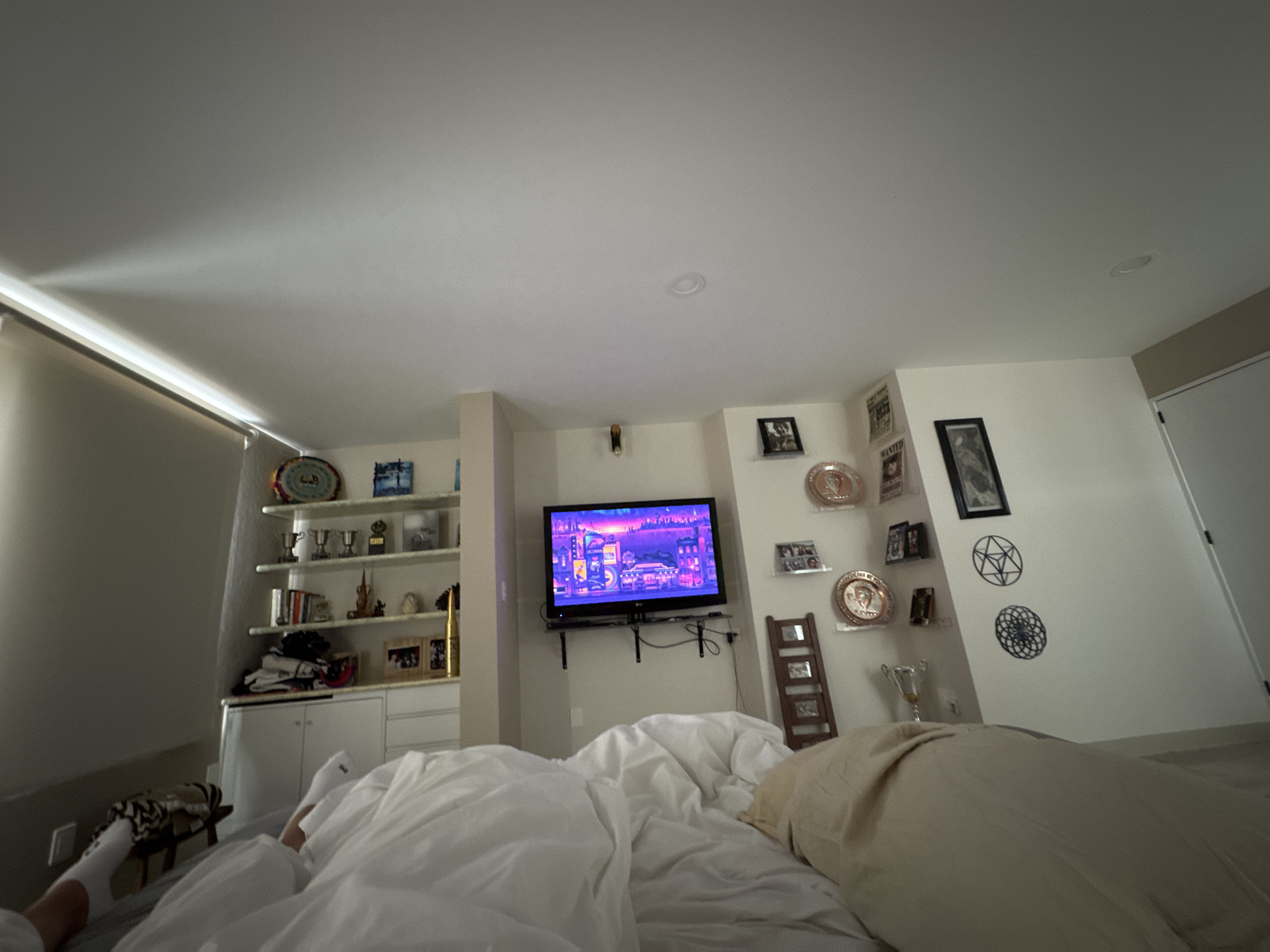 The image shows a bedroom with a bed in the foreground. The bed is covered in white and beige bedding. A person's foot, wearing a white sock, is visible on the left side of the bed. A TV is mounted on the wall between two built-in shelves with various decorative items. The room appears to be well-lit. The location is not possible to determine from this image.