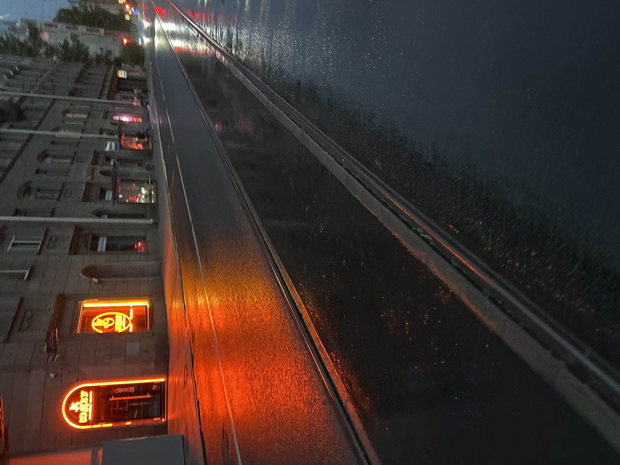 The image captures a street scene at dusk, likely after rainfall, in an urban environment. The foreground is dominated by a wet asphalt road with visible tram tracks, reflecting the ambient light. To the left, a building facade is illuminated by a bright, arched neon sign with text in an unfamiliar script, casting a strong orange glow onto the wet pavement. Next to this entrance, a storefront window also displays a similar neon circular logo, further contributing to the warm lighting. Further down the street, more buildings with windows line the road, and the taillights of a passing vehicle are visible, along with reflections on the wet road surface. The overall atmosphere is moody and atmospheric, suggesting an evening in a city with active nightlife or commercial activity.