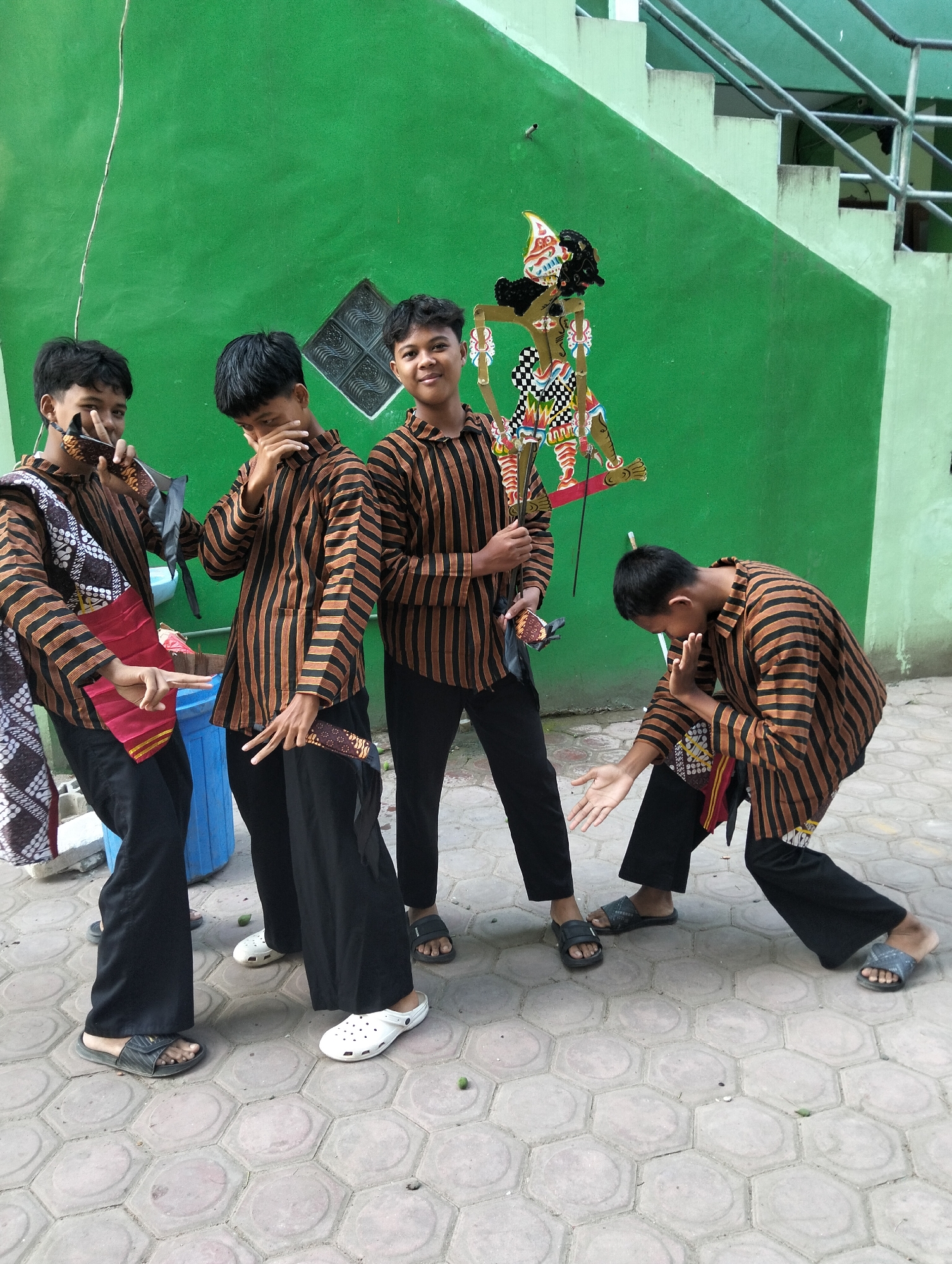 Four young males, appearing to be teenagers or young adults, are posing for a photograph in front of a bright green wall in what is identified as Kranji, Indonesia. All four individuals are wearing matching black pants and brown and black striped long-sleeved shirts.
On the left, two individuals are positioned side-by-side. The leftmost individual is holding a traditional Javanese wayang puppet with a pointed headdress and dark hair. They are also holding a folded red and yellow cloth. This individual is looking towards the camera and has their right hand raised, holding the puppet. The second individual from the left is also looking towards the camera, with their left hand covering their mouth and their right hand resting on their chin.
In the center, a third individual, dressed identically, is holding a similar wayang puppet higher up and looking towards the camera with a slight smile. Their right hand is gesturing towards the puppet.
On the right, the fourth individual is crouched down with their right arm extended and their palm facing forward. Their left arm is bent at the elbow, with their hand near their chest. This individual is looking downwards and to their left.
The ground is paved with hexagonal concrete tiles. Behind the individuals, the green wall has a diamond-shaped patterned grate. To the right of the green wall, a light-colored staircase with a metal railing is visible, suggesting an outdoor or semi-outdoor setting. A blue plastic bin is positioned behind the two individuals on the left. The overall scene appears to be a candid or posed moment capturing the individuals and their cultural props.