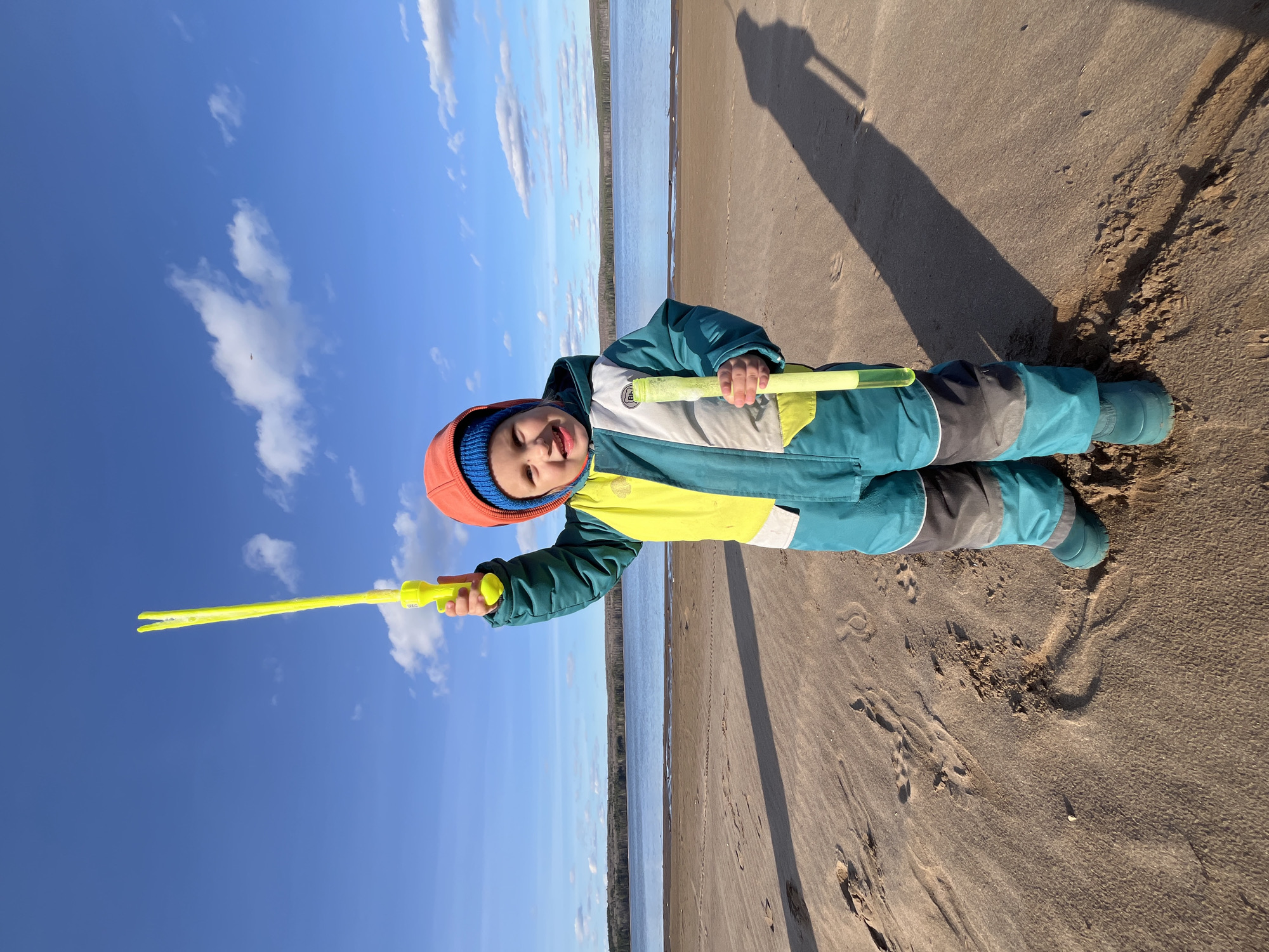 A young child, dressed in an orange hat with a blue trim, a teal, yellow, white, and grey snowsuit, and teal boots, stands on a sandy beach. The child is holding a long yellow plastic toy. The background includes a blue sky with scattered white clouds, a body of water, and a line of trees on the opposite shore. The child's shadow is cast on the sand.
The location is Chaikovsky, Russia.