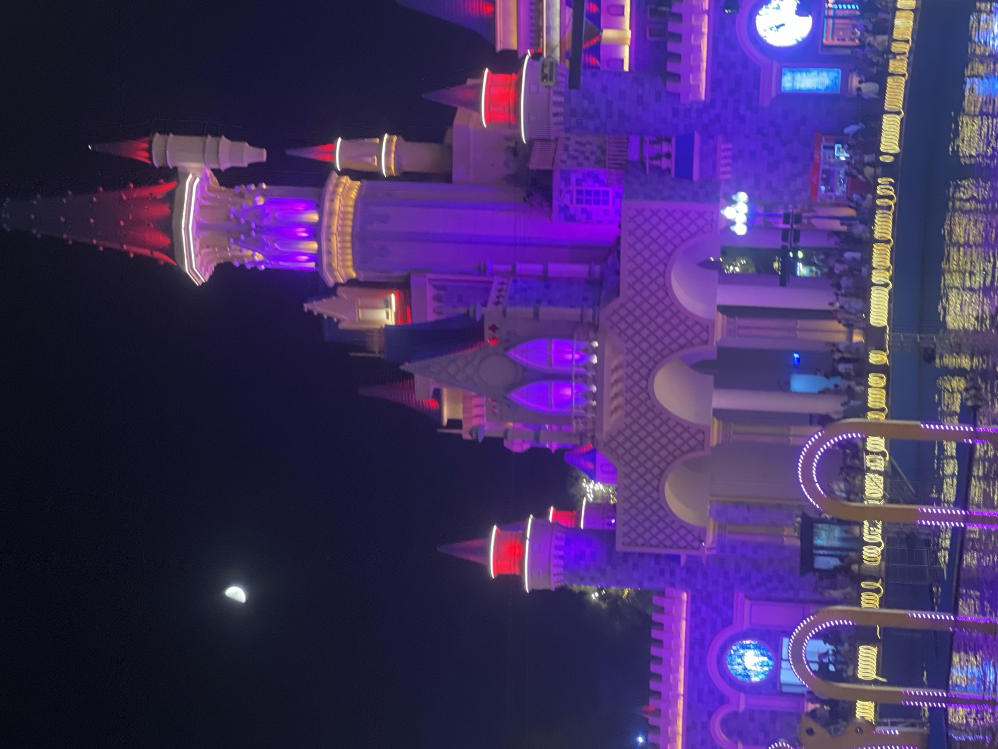 The image depicts a brightly lit, fairy-tale-like castle at night. The castle is illuminated with vibrant purple and red lights, accentuating its turrets and architectural details. A sliver of the moon is visible in the dark night sky. In the foreground, there appears to be a body of water reflecting the castle's lights, and a crowd of people can be seen gathered in front of the structure, suggesting it is a public attraction or event. The overall scene evokes a sense of fantasy and spectacle. The location is described as Unknown, Unknown.