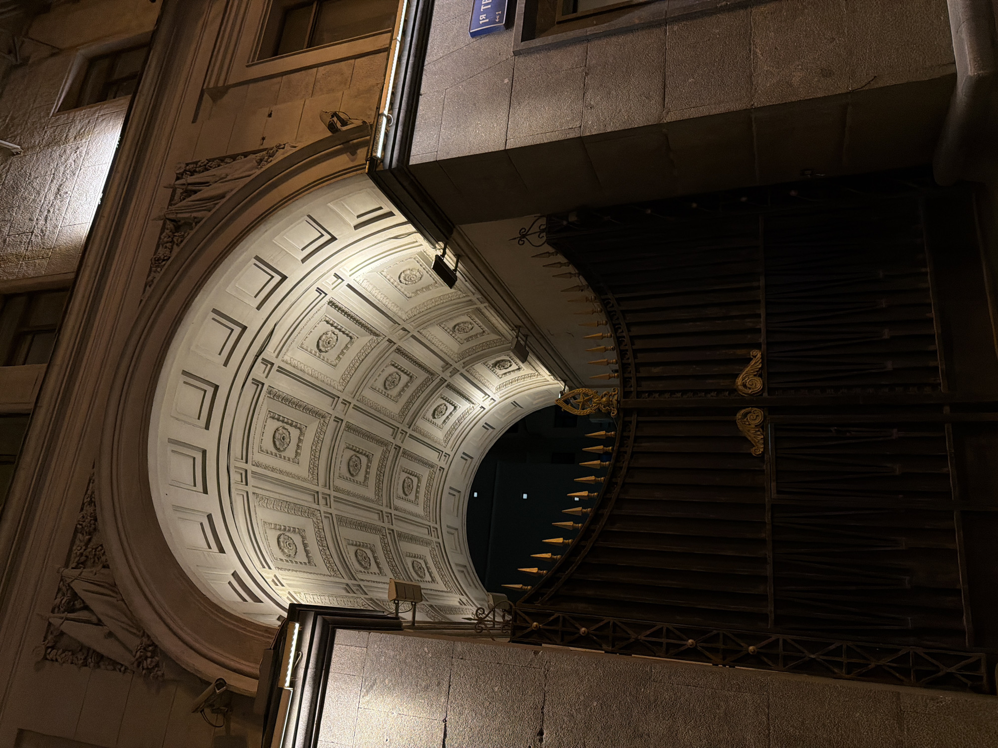 The image captures an exterior view of a building at night in Moscow, Russia. An elaborate archway with ornate, classical detailing dominates the left side of the frame, featuring a coffered ceiling pattern with decorative rosettes and nested square designs. The archway is brightly illuminated, contrasting with the darker right side of the image where a large, dark iron gate with pointed finials and decorative scrollwork is visible. A street sign reading "1-й Твер" is affixed to the building above the archway, providing a geographical reference. The building facade is constructed of a light-colored stone, possibly granite or limestone, exhibiting a rough texture. The overall scene conveys a sense of historical architecture illuminated in the urban environment.