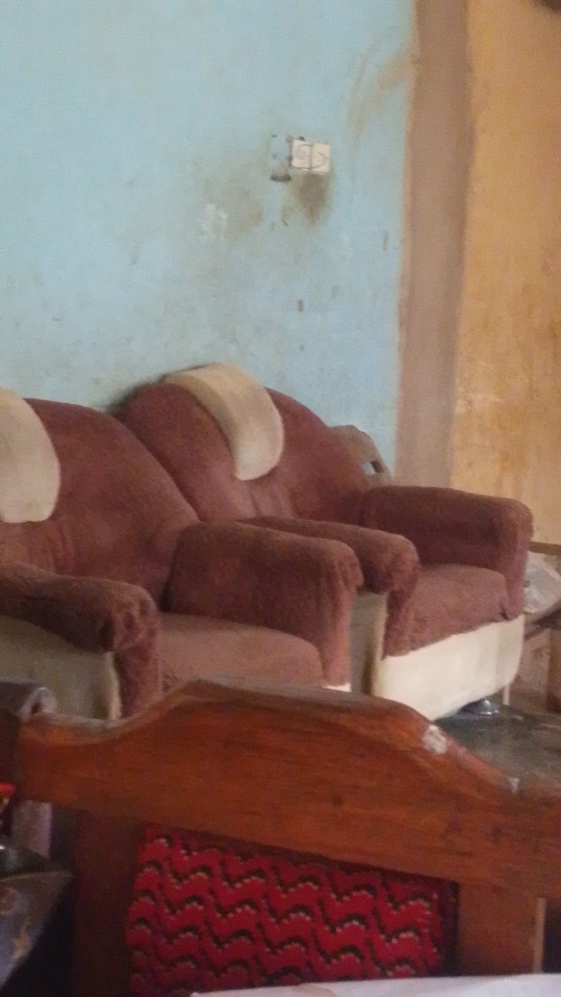 The image depicts two comfortable armchairs with maroon upholstery, placed side-by-side in a room. The armchairs have light-colored headrests. In the foreground, a wooden furniture piece with a red, zig-zag patterned cushion is visible. The wall behind the armchairs is painted light blue and shows some stains. There are electrical outlets on the wall. The room appears to be indoors, with warm lighting, suggesting it is daytime. The setting suggests a domestic environment in Donga, Nigeria. No people or specific activities are visible in the image.