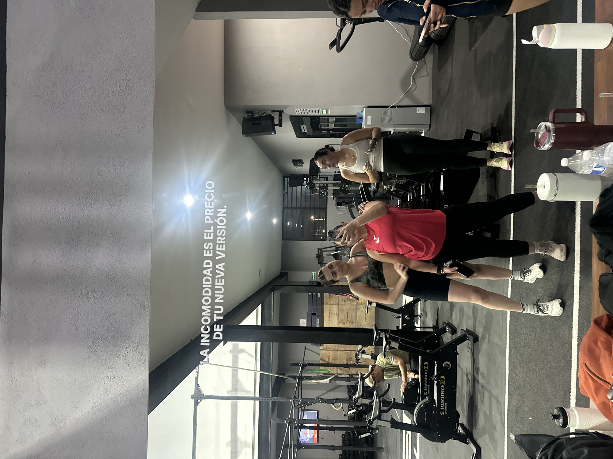 The image shows a group of women at a gym in San Bernardino Tlaxcalancingo, Mexico. Some are on stationary bikes, others are standing and taking a selfie. Workout equipment is visible in the background. There's also a motivational message written on the wall, with water bottles on the floor. The gym setting suggests the people are engaged in fitness activities.