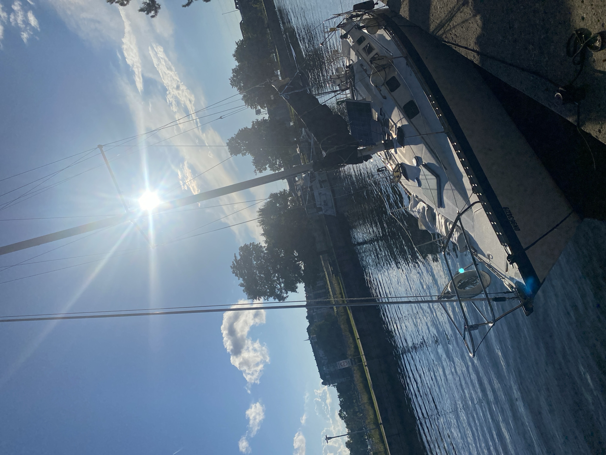 A sailboat is docked on a concrete pier under a bright, sunlit sky. The boat, with a dark sail furled, is tilted slightly towards the water. The water's surface is rippled and reflects the sunlight. In the background, trees line the far bank of the canal, and beyond them, a large, multi-story building is visible. The boat's registration number, "ON963412B", is visible on its hull. The scene is located in Sainte-Anne-de-Bellevue, Canada.