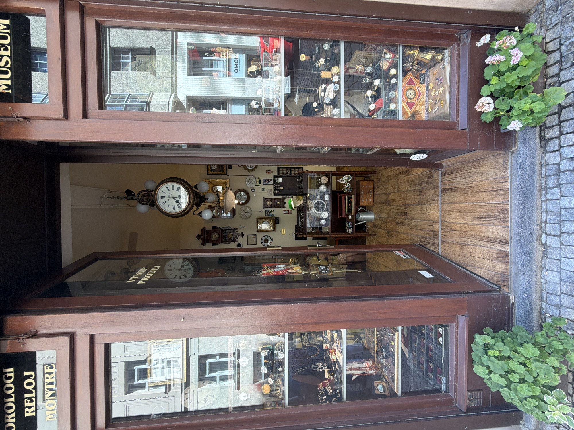 The image shows the exterior of a clock and watch shop in Prague, Czechia, identified by the signage "OROLOGI RELOJ MONTRE MUSEUM". The shop has a large wooden door that is open, revealing the interior. The storefront is filled with display cases showcasing numerous clocks and watches of various styles and ages. On the left side, a display case contains jewelry and watches. Inside the open doorway, a large ornate clock hangs from the ceiling. Further inside, more clocks and display shelves are visible, along with a person partially obscured behind displays. The floor inside the shop is made of light-colored wood planks, and the exterior pavement is cobblestone. Two planters with green and pink flowers are situated in front of the shop entrance. Reflections of buildings across the street are visible in the shop windows.