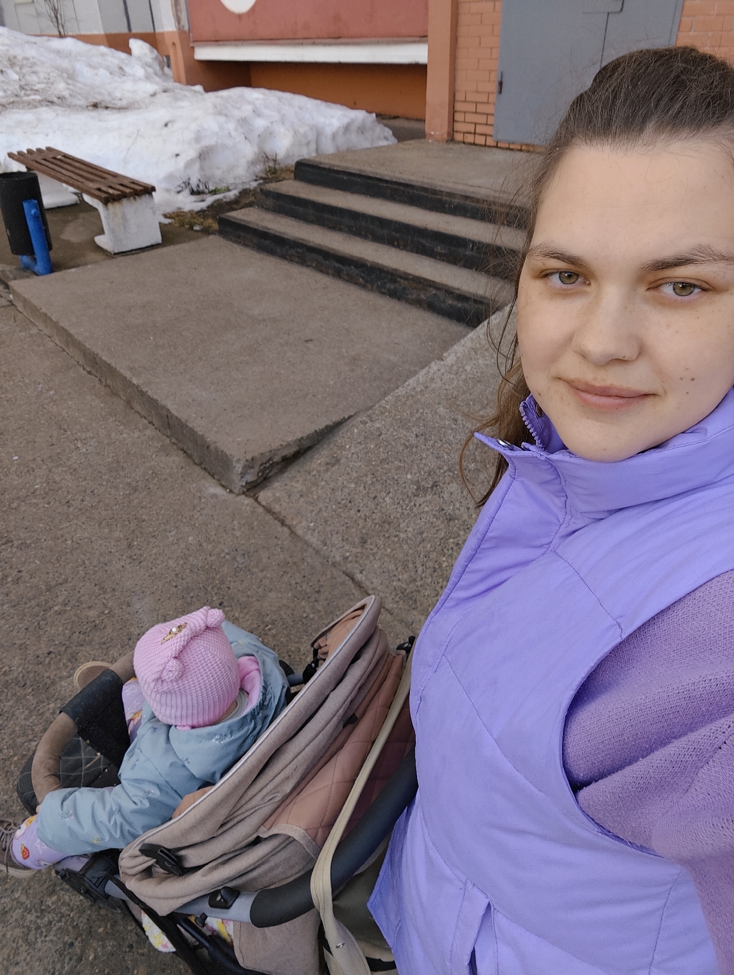 The image shows a woman taking a selfie with a baby in a stroller.
The woman is light-skinned with brown hair and eyes. She is wearing a light purple vest over a purple sweater. The baby in the stroller is bundled in a blue snowsuit and a pink knit hat.
The background shows a building with red brick, concrete steps, a bench and a snow pile. The location is the city of Chaikovsky, Russia.
