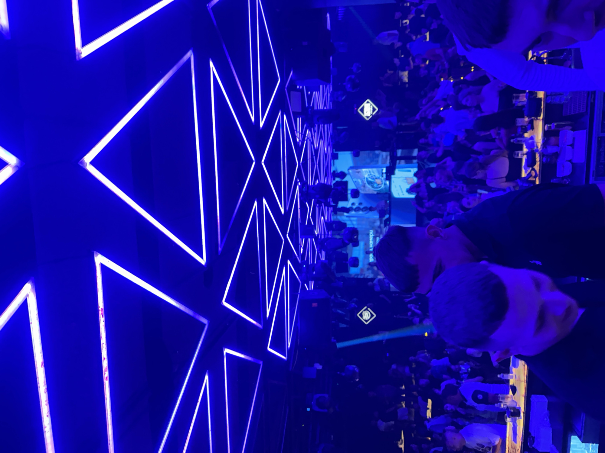 The image depicts the interior of a nightclub in Ruse, Bulgaria, illuminated by vibrant blue neon lights forming triangular geometric patterns across the ceiling. The club appears to be crowded, with numerous patrons visible in the background, some standing and others seated at tables. In the foreground, three individuals are partially visible. Two men, with closely cropped hair, are the most prominent subjects. One is looking down and to the right, while the other is positioned behind and slightly to the left of the first. Another man, seen from the shoulders up, is in the far right of the frame. The environment suggests a lively atmosphere, typical of a nightclub setting, with lighting playing a significant role in the ambiance.