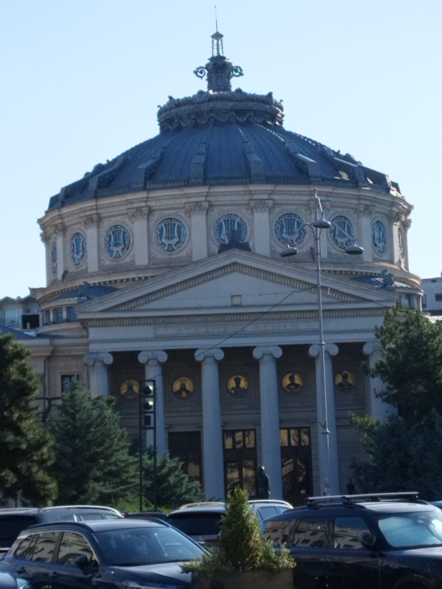 The image shows the Romanian Athenaeum, a concert hall in Bucharest, Romania. The building features a prominent dome, classical columns, and decorative circular motifs. In the foreground, several cars are parked, suggesting an urban environment. Trees are visible on the left and right sides of the building, and a street light with multiple lamps stands in front of the Athenaeum. The sky is clear and blue.