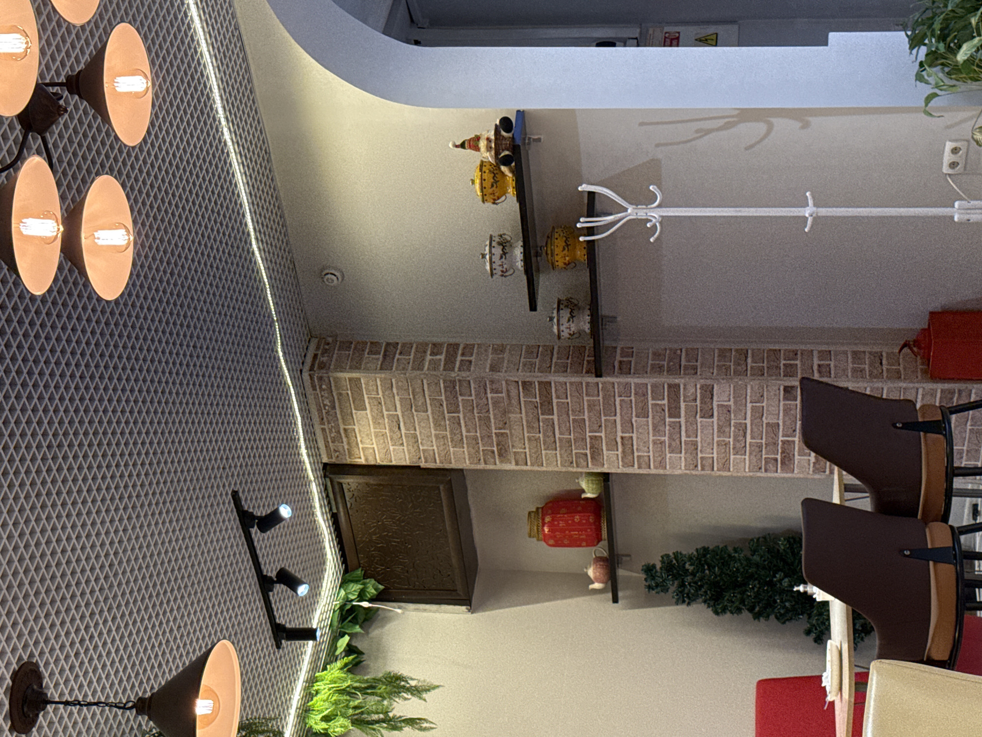 The image depicts an interior scene, likely a cafe or restaurant, with a focus on decorative elements and seating. The ceiling features a textured grid pattern with recessed lighting and several industrial-style pendant lamps with exposed bulbs. Track lighting is also visible on the ceiling.
On the wall, there are shelves displaying decorative items. On a higher shelf, a dark wooden decorative panel is mounted. Below that, on a black shelf, sits a large red cylindrical vase with intricate gold detailing, flanked by two small teapot-like objects, one light pink and one pale green. A small, artificial evergreen tree is placed on a lower shelf.
Further to the right, two more black shelves are visible. The upper shelf holds three decorative ceramic jars with lids: two white with blue patterns and one yellow. A small festive figure, possibly a gnome or Santa Claus, wearing a hat, is perched on the far right of this shelf. The lower shelf has two yellow decorative jars with lids and a white coat rack with multiple hooks stands prominently in the foreground to the right.
In the lower left portion of the image, a table with chairs is partially visible. Two dark brown chairs with brown upholstery and black metal frames are in focus, positioned around a light-colored table. A white plate with a small amount of food is on the table. A red chair is partially visible behind the table.
The walls are painted a neutral, light color, with a section of exposed brickwork forming a column to the right of the shelves. An arched doorway is visible on the far right of the image, leading to another area. A power outlet is visible on the wall near the base of the coat rack. A red fire extinguisher is located on the floor to the left of the brick column. A potted plant is visible in the lower right corner.
There are no people present in the image. The overall atmosphere suggests a cozy and perhaps themed establishment. This image was captured in Krasnoyarsk, Russia.
